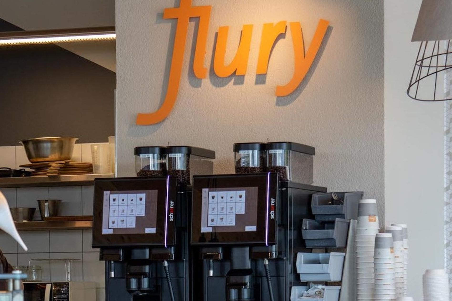 Two coffee machines beneath an orange "flury" sign in a modern café setting, with stacked cups and a shelf in the background.