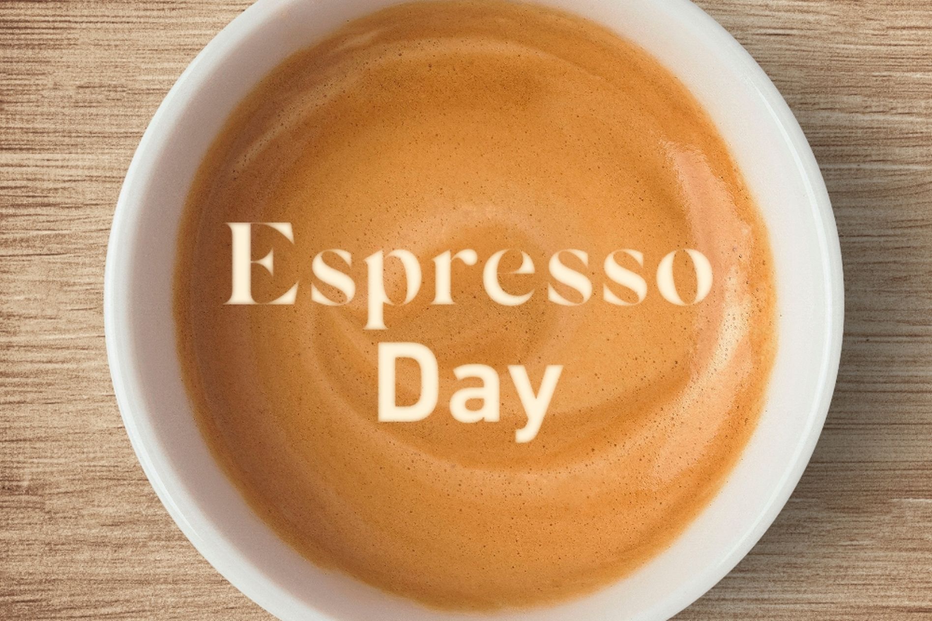 A cup of espresso with a creamy swirl, viewed from above, on a wooden surface. Text overlay reads "Espresso Day."