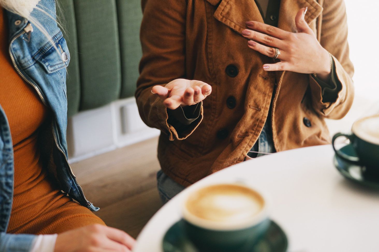 Two people sitting at a table with coffee cups, engaged in conversation. One gestures with hands, wearing a brown jacket.