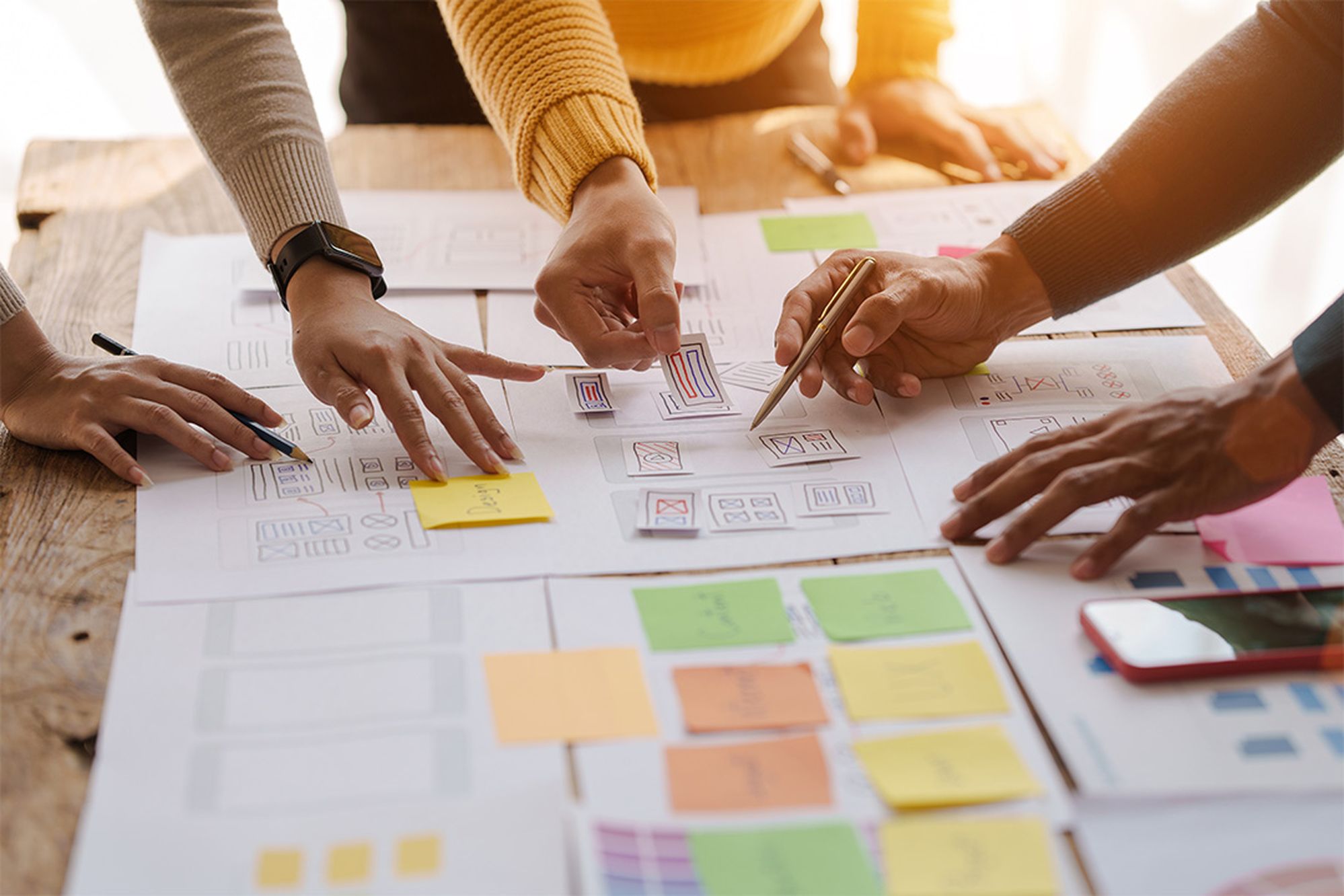 Hands of diverse individuals collaborating on design sketches and notes spread on a wooden table, with colorful sticky notes and a smartphone nearby.