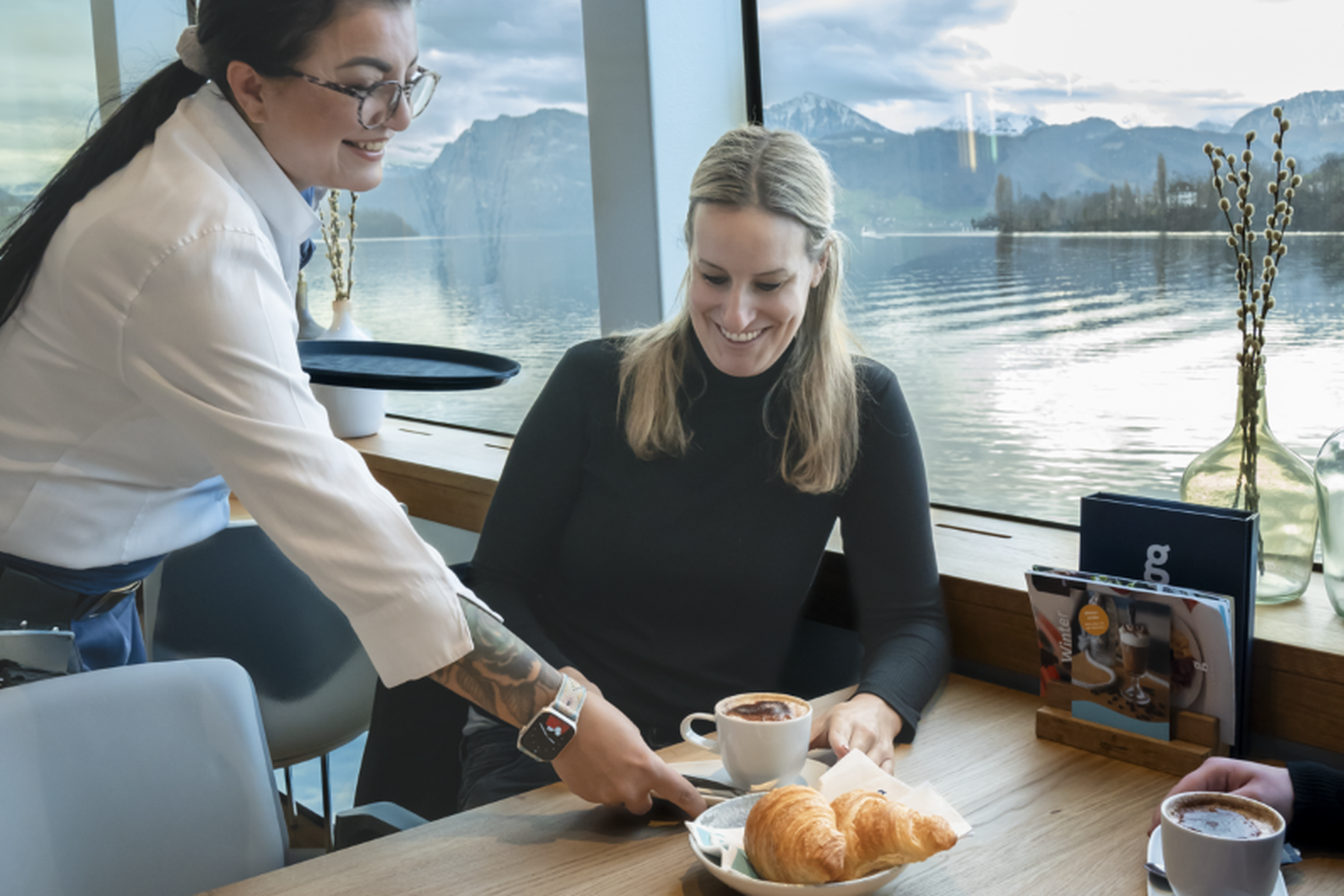 A woman in a cafe receives coffee and croissants from a server. The setting overlooks a lake with mountains in the background.