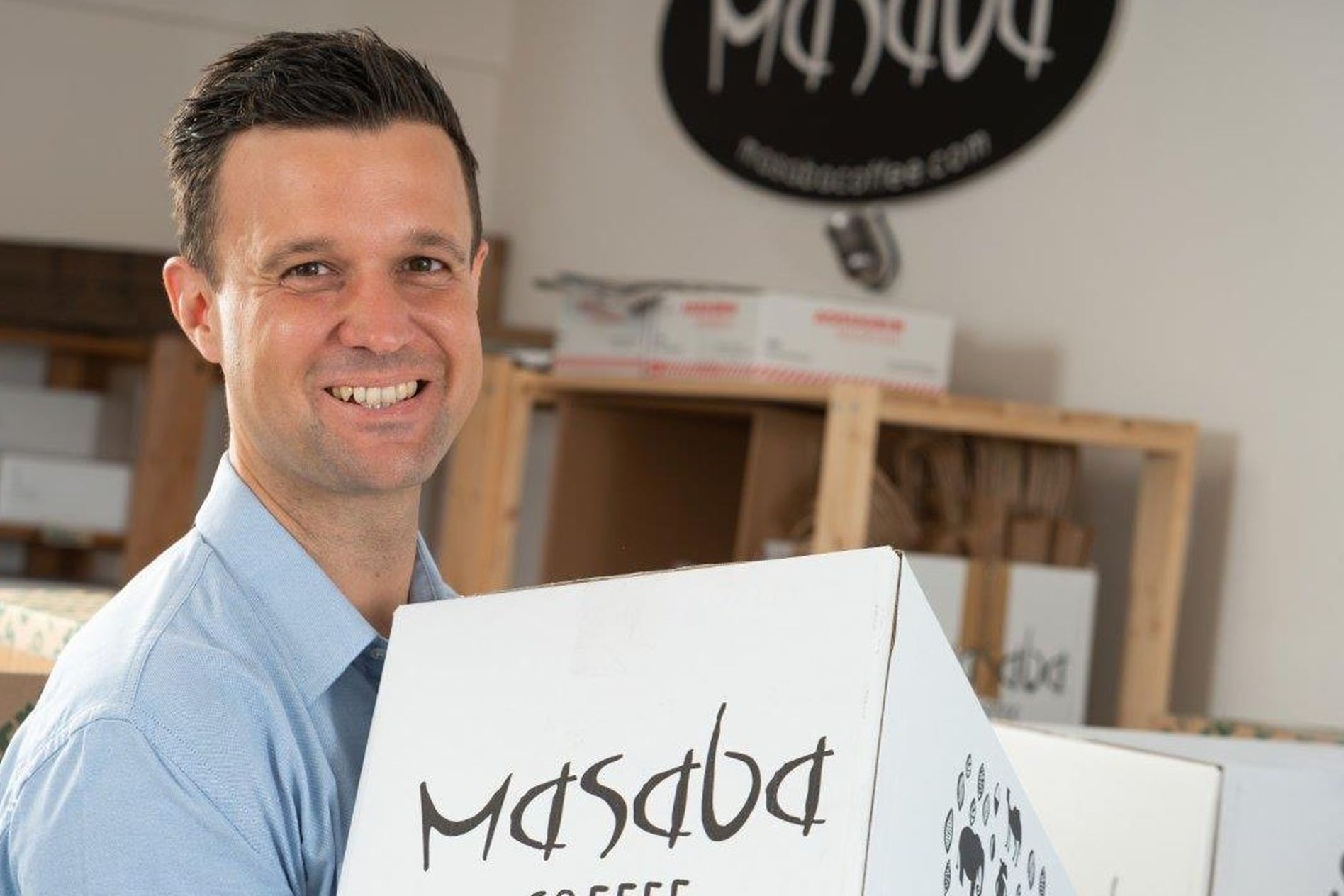 Smiling person holding a white box labeled "Masaba Coffee" in a room with more boxes and shelves in the background.