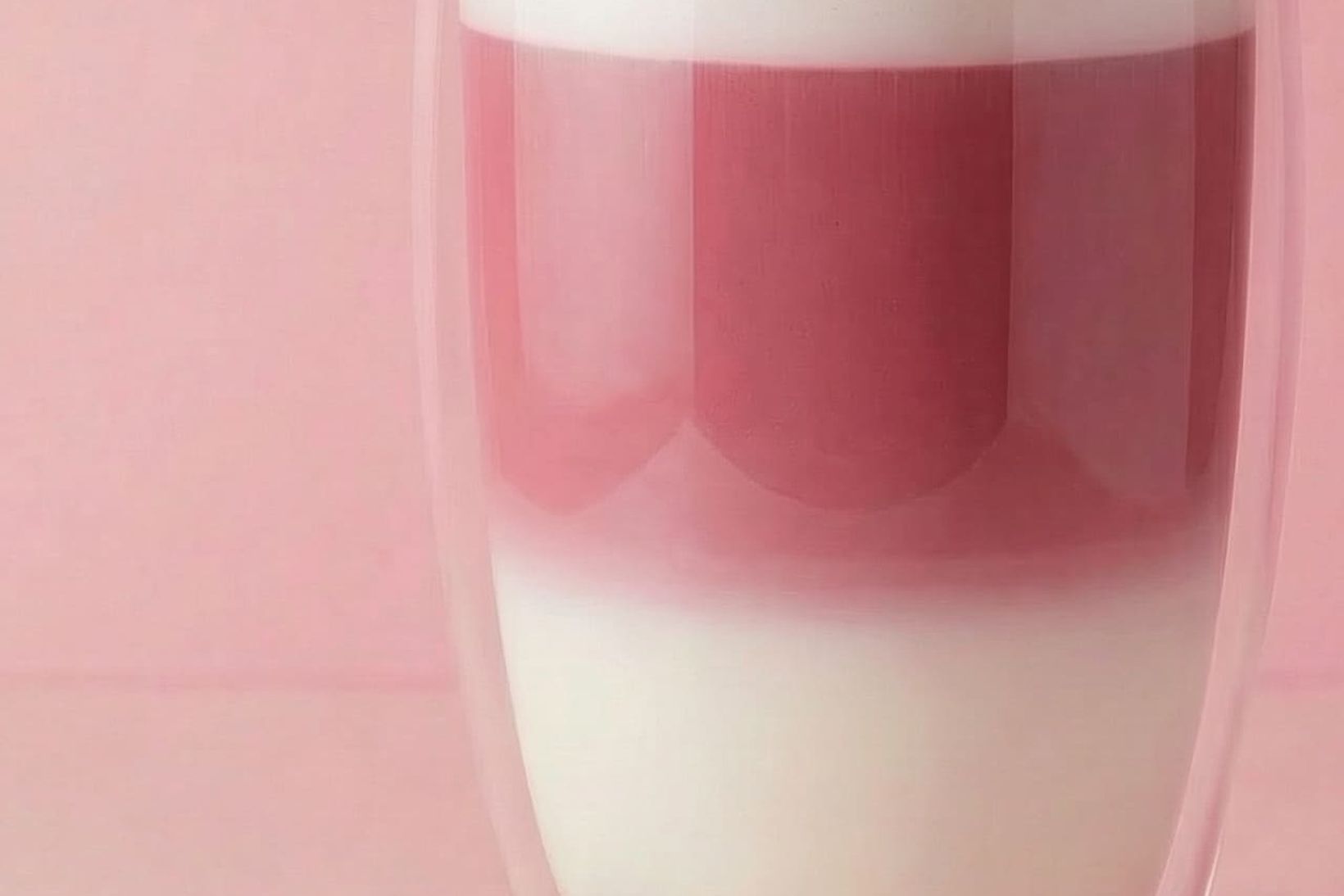 A double-walled glass filled with layered pink and white latte-style drink, next to a small mound of pink powder, against a pink background.