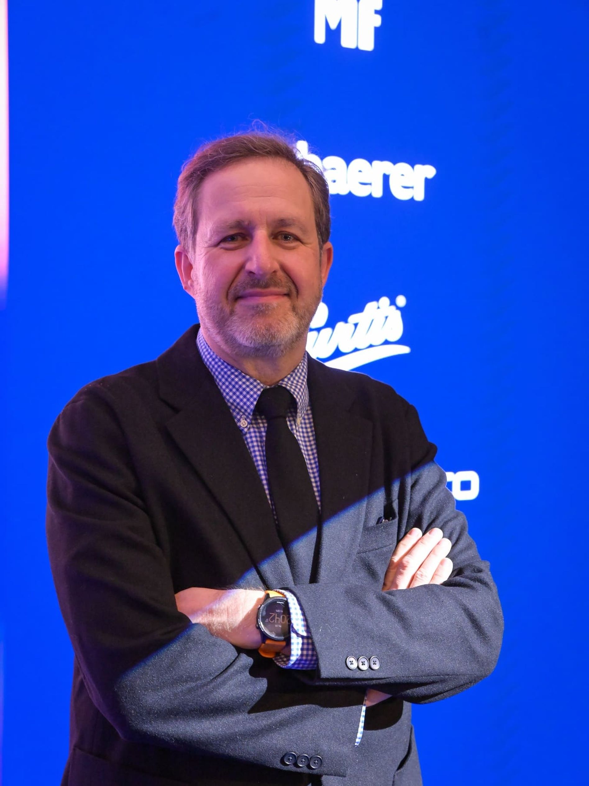 Person in a suit with arms crossed, standing in front of a blue background with industry-related logos.