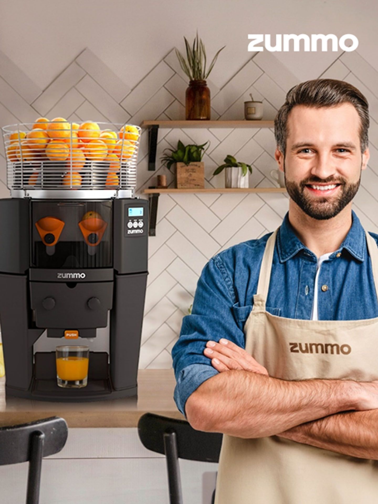 A smiling person wearing a Zummo apron stands beside a Zummo juicer filled with oranges on a kitchen counter.