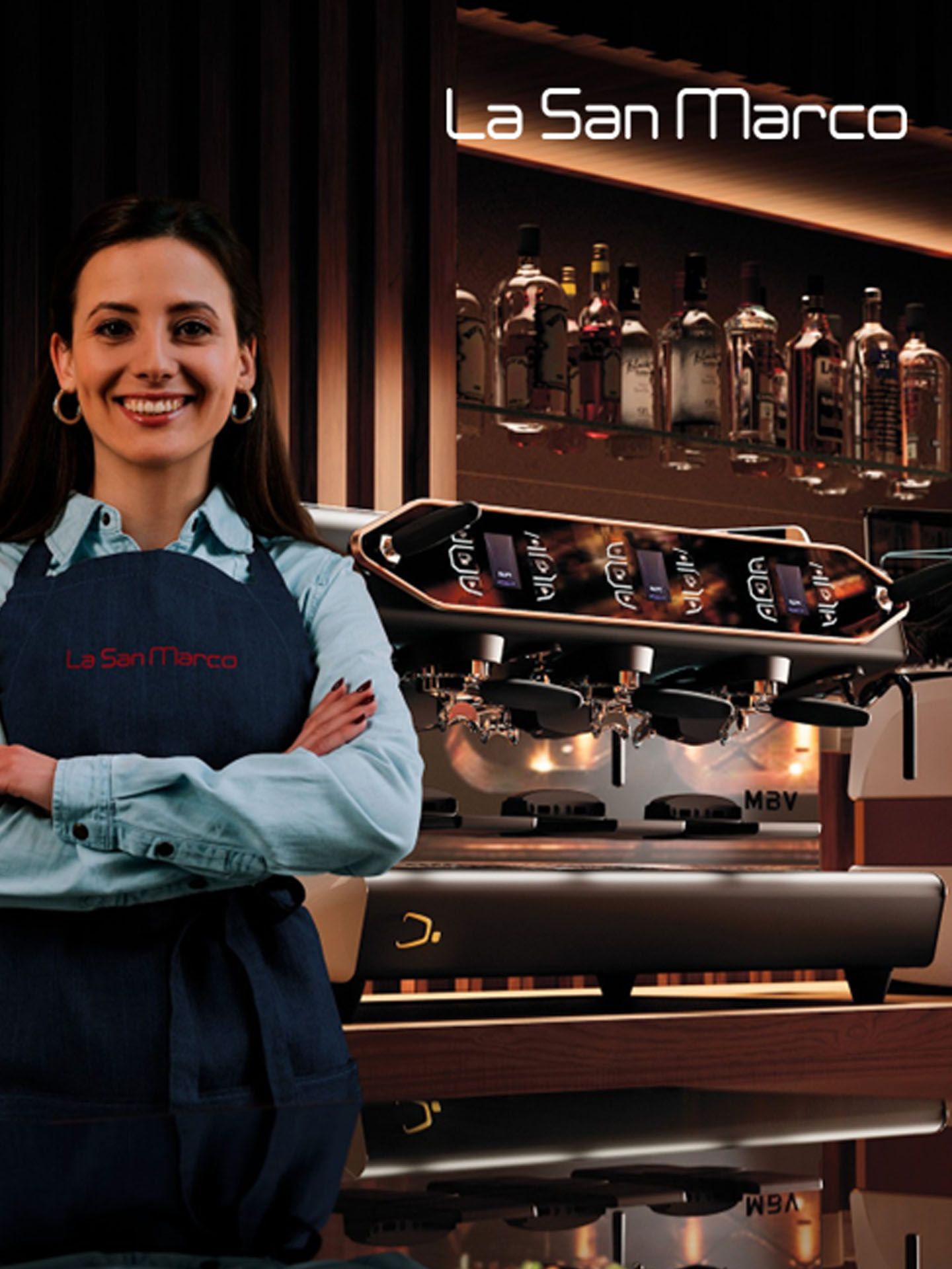 Smiling barista in apron stands in front of La San Marco coffee machine, with bottles displayed on a shelf in the background.