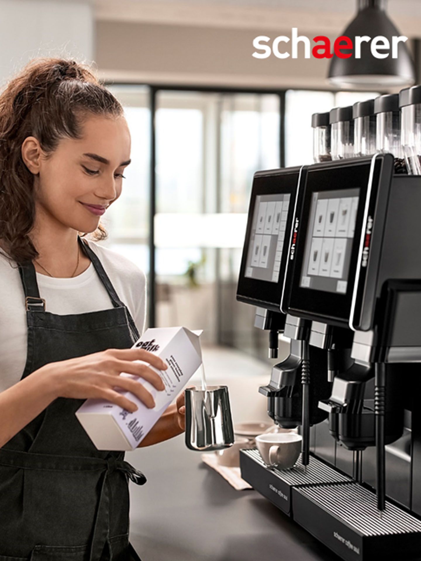 Barista pouring milk into a frothing pitcher next to a Schaerer coffee machine with touchscreens.