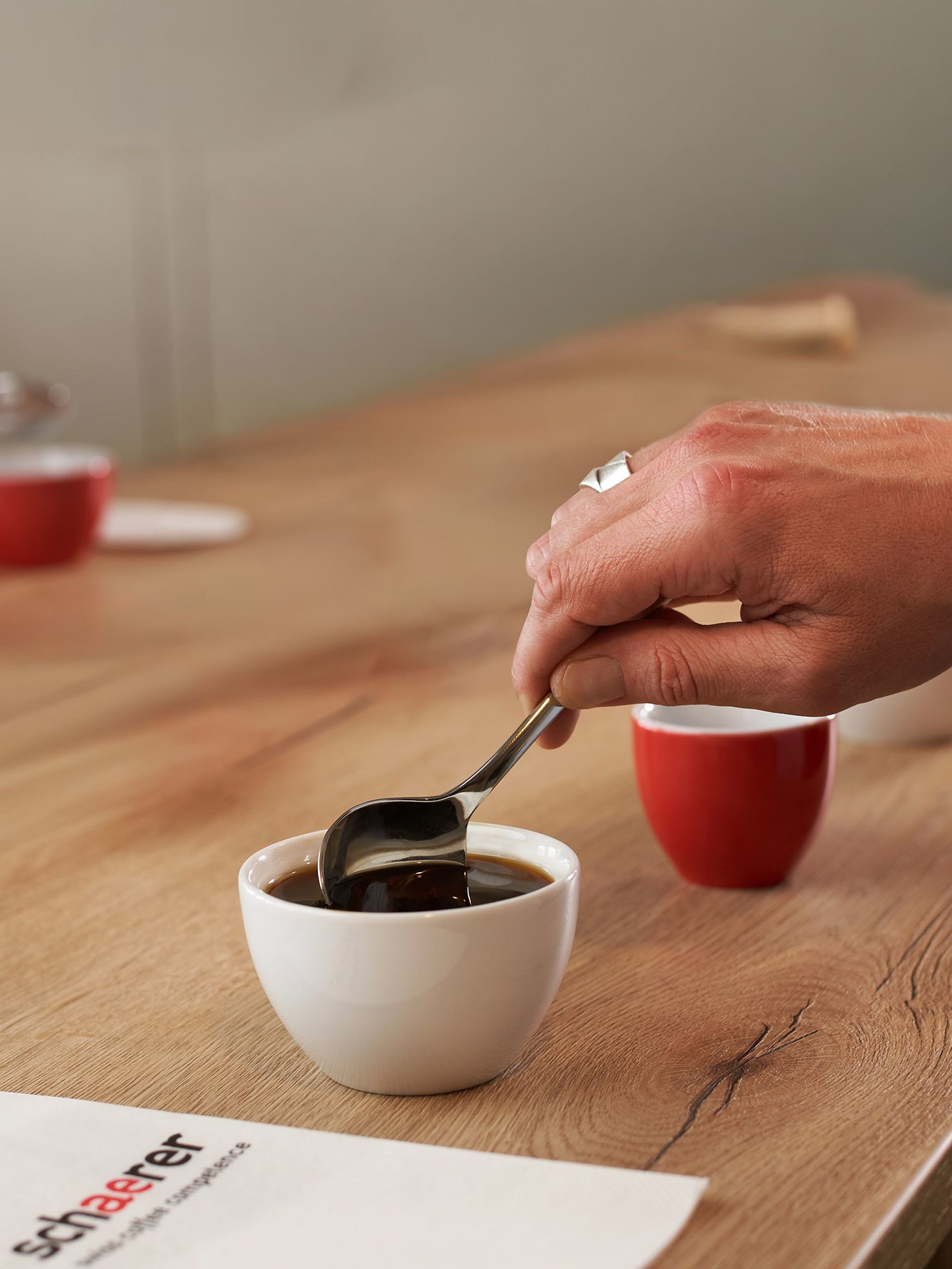 A hand holding a spoon stirs coffee in a white cup on a wooden table, surrounded by other cups and a metal kettle in the background.