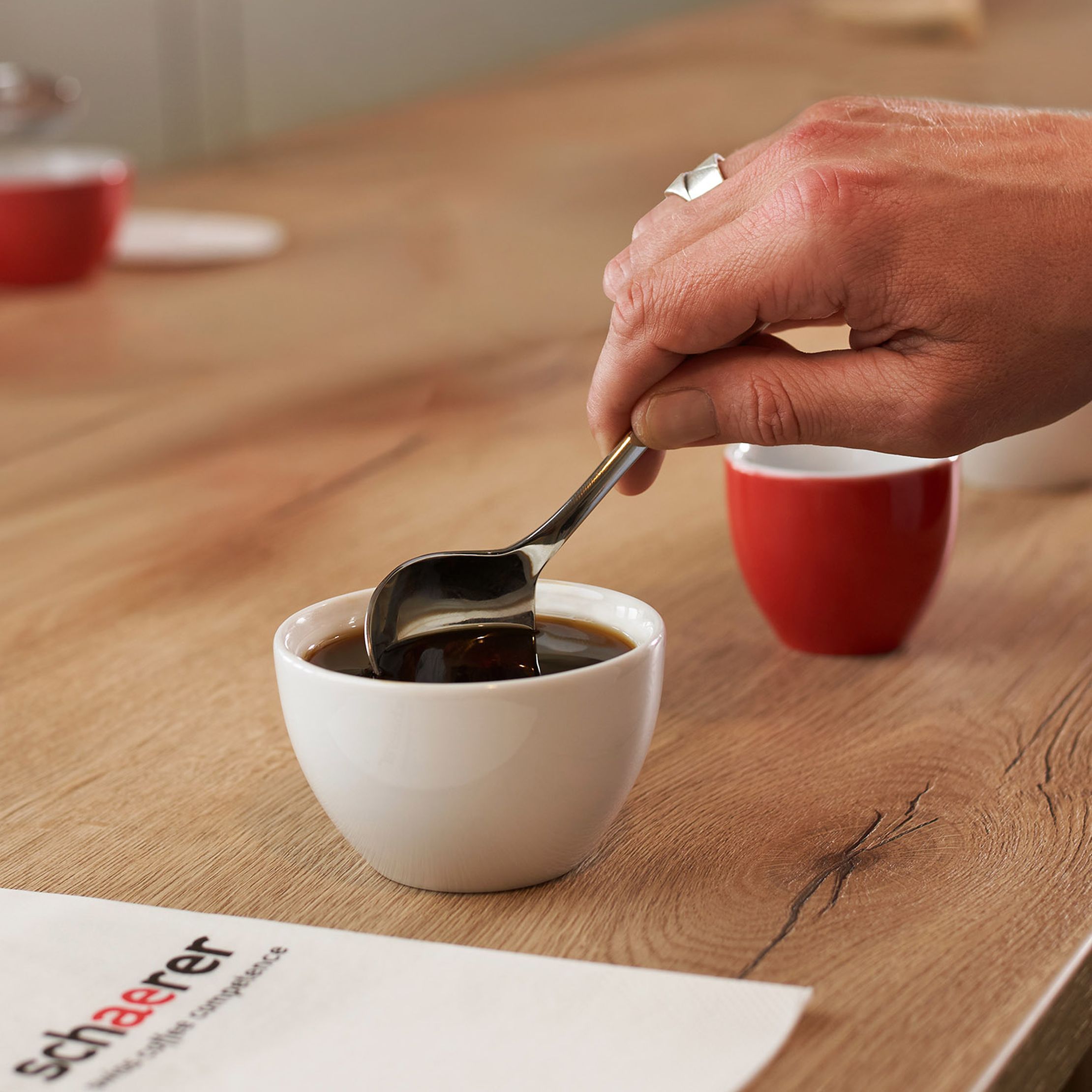 A hand holding a spoon stirs coffee in a white cup for cupping