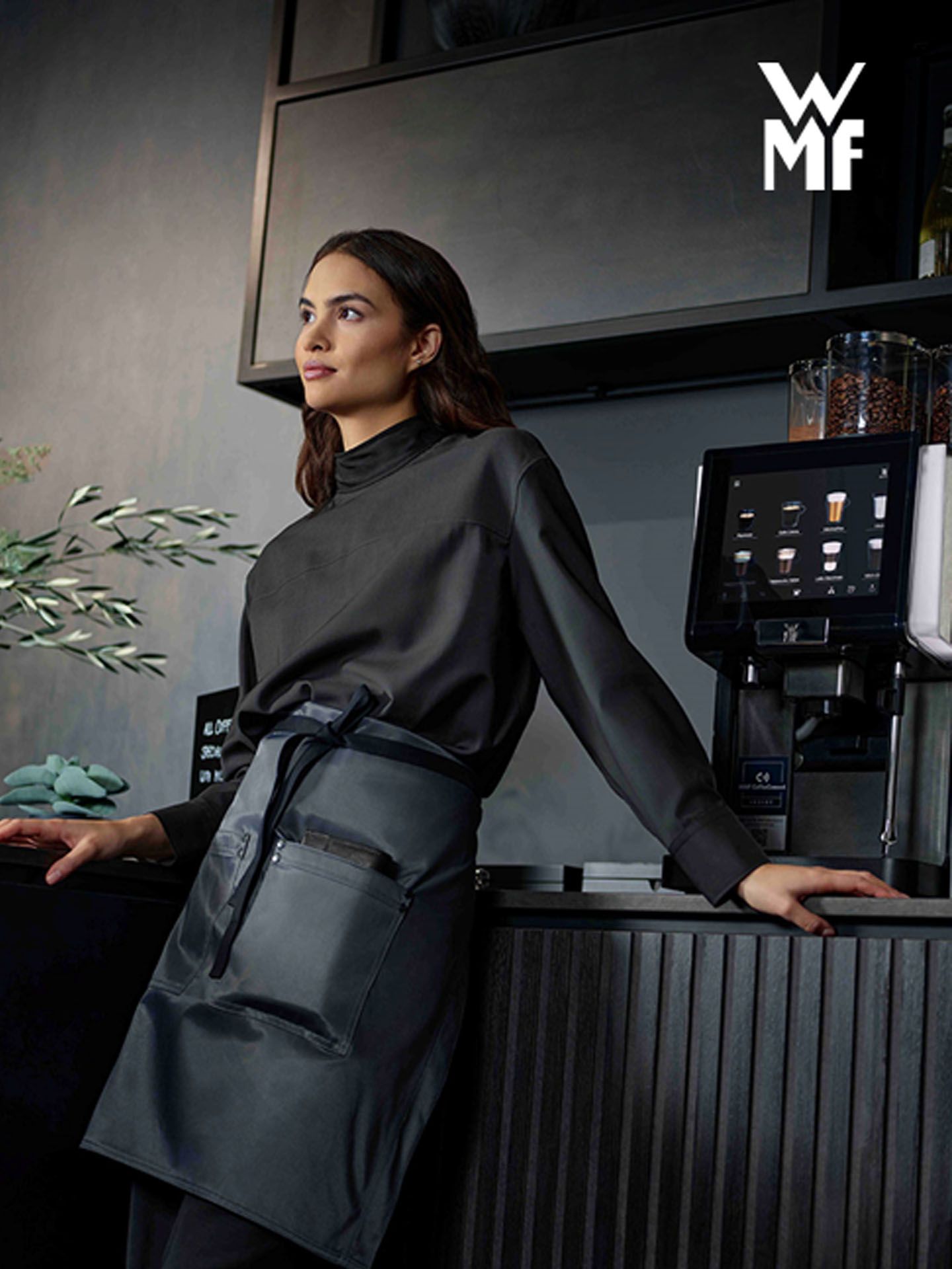Woman in a dark uniform leans on a counter beside a WMF coffee machine in a modern café setting.