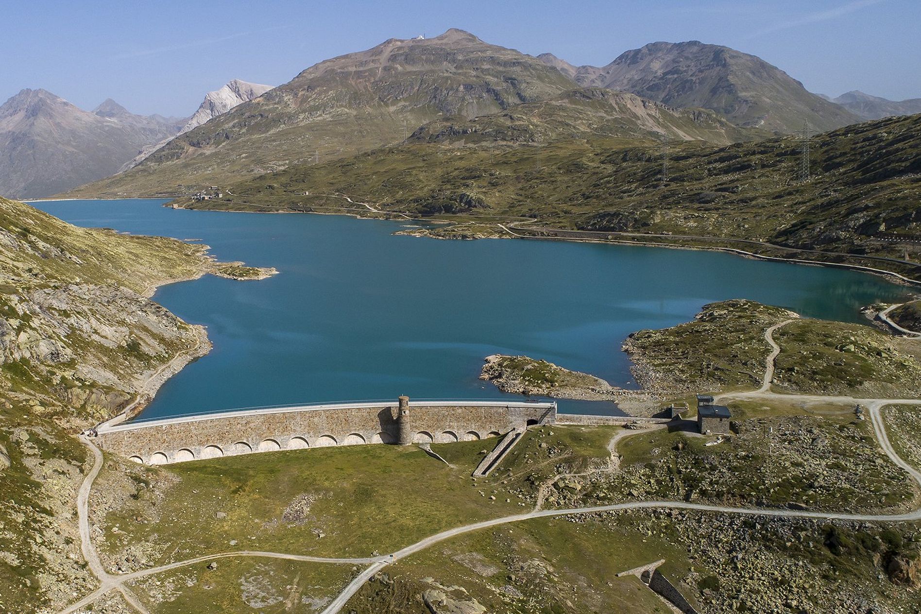 Aerial view of a large dam and reservoir surrounded by mountainous terrain under a clear blue sky.