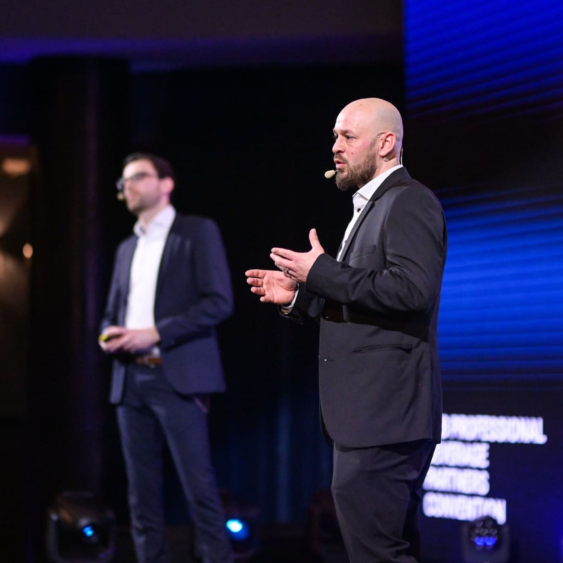 Two men in suits are speaking on a stage with a lit background. One man is gesturing while addressing an audience.