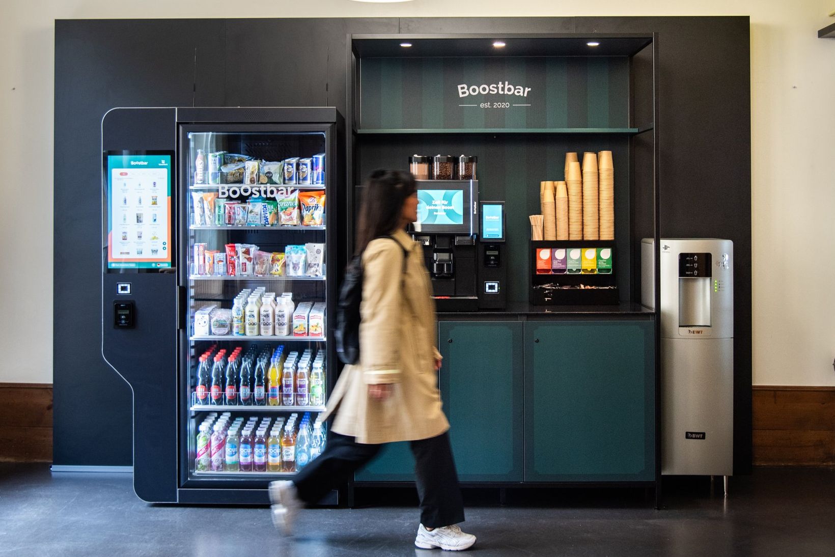 A person walks past a vending machine and coffee station labeled "Boostbar," featuring snacks, drinks, and a coffee dispenser.