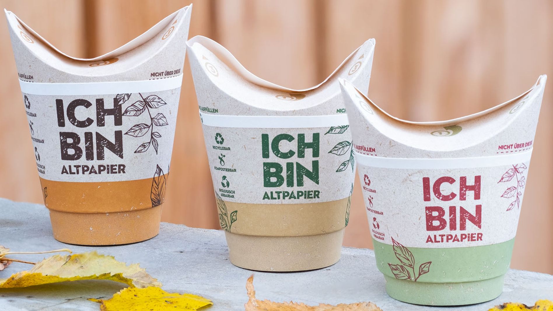 Three eco-friendly paper cups with "ICH BIN ALTPAPIER" text, in different colors, placed on a ledge with scattered autumn leaves.
