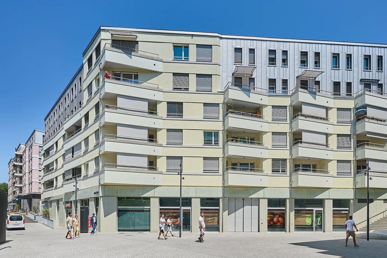 Modern multi-story apartment building with balconies, ground-floor shops, and pedestrians walking on a sunny day.