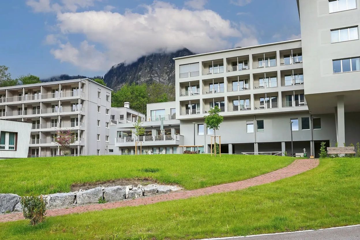 Modern apartment buildings with balconies, set against a mountainous backdrop, with a green lawn and a winding brick path in the foreground.