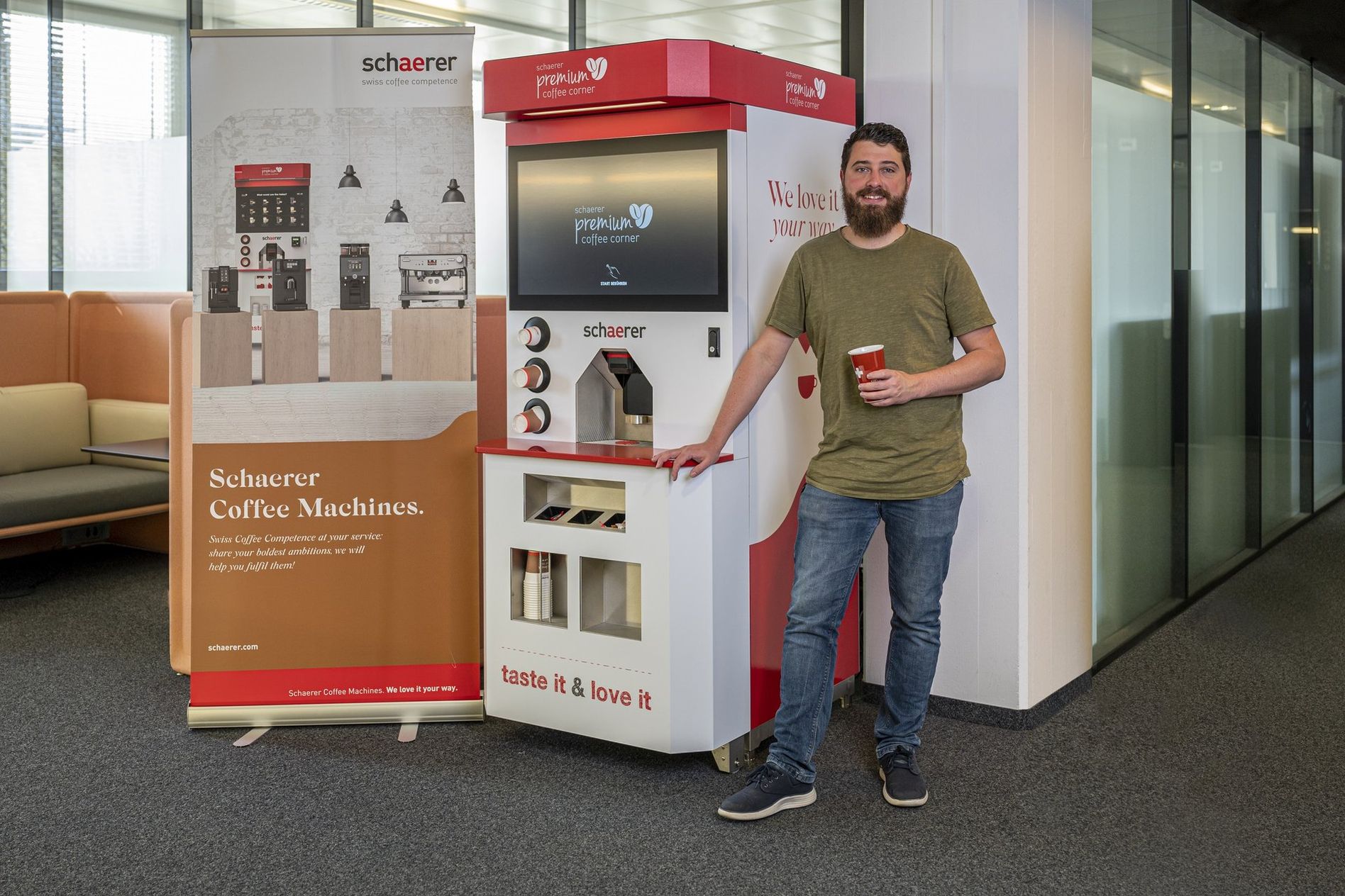 Man holding a coffee cup stands beside a Schaerer coffee machine in an office setting, with a promotional banner in the background.