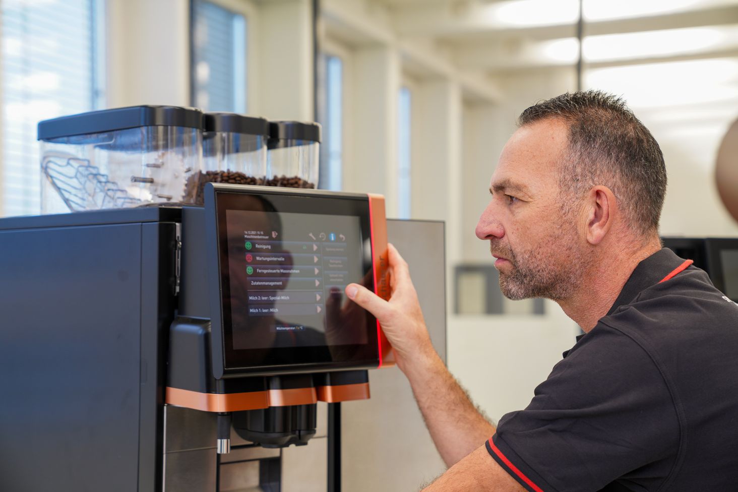 A man operates a touchscreen coffee machine, adjusting settings. The machine has visible coffee bean containers on top.