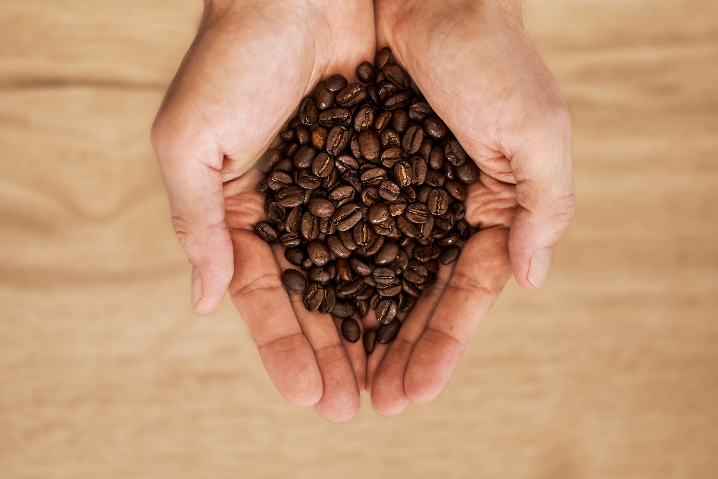 Hands holding a pile of roasted coffee beans over a wooden surface.