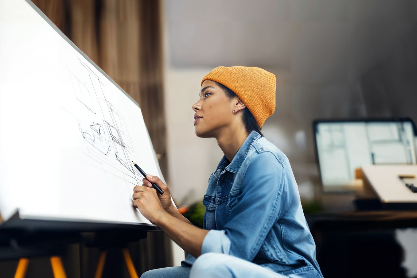 Person in a yellow beanie sketching on a large paper at an easel, focused and seated, with a blurred background of a room and a computer.