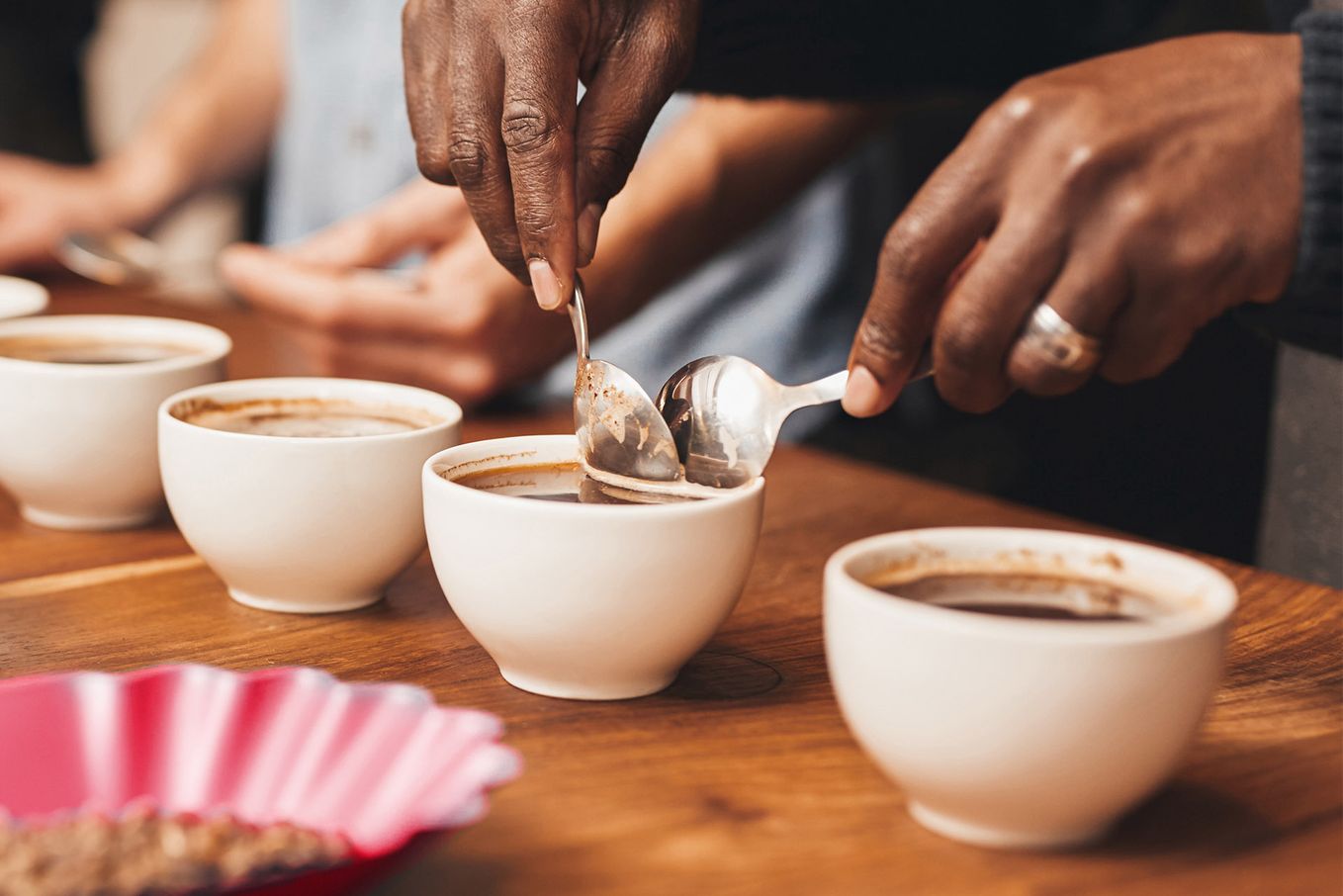 Hands holding spoons over white cups filled with coffee on a wooden table, with a pink coffee filter nearby.