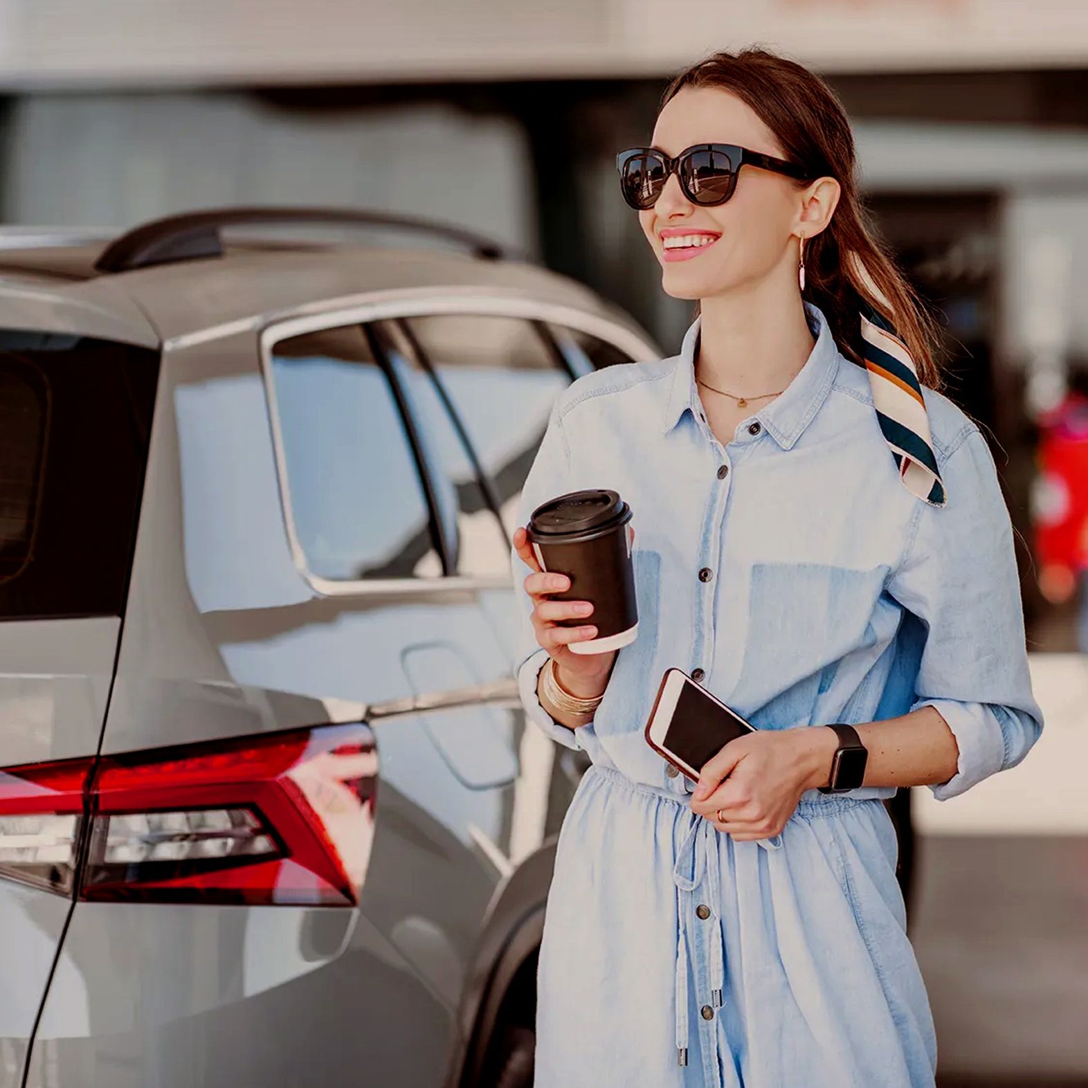 Woman in denim dress holding coffee and phone, standing by a car, wearing sunglasses and a scarf in her hair, smiling.
