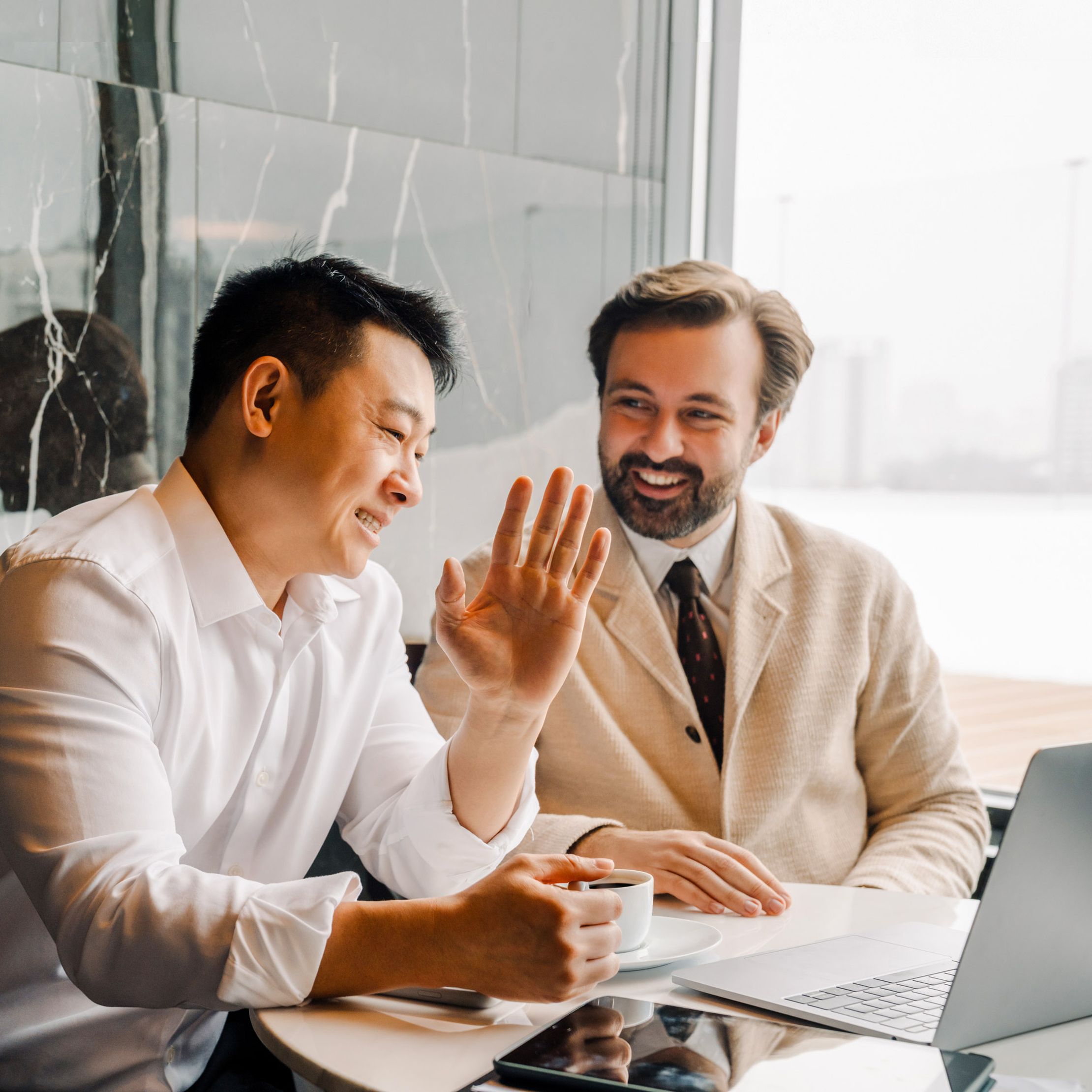 Two men in business attire sit at a table with a laptop, smiling and conversing. One gestures while holding a pen, and a cup of coffee is on the table.