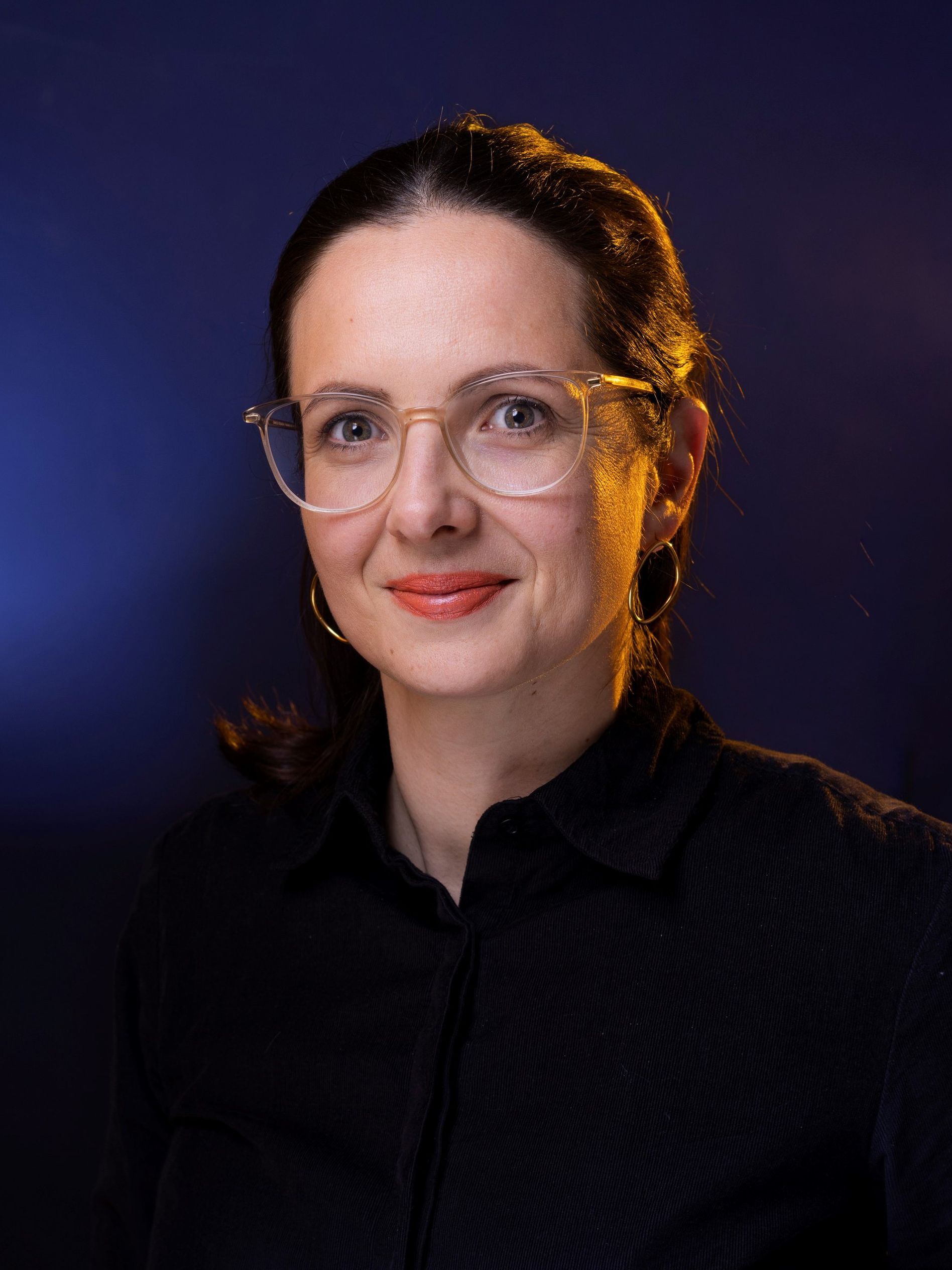 Woman with glasses and hoop earrings, wearing a black shirt, smiles slightly against a dark blue background.