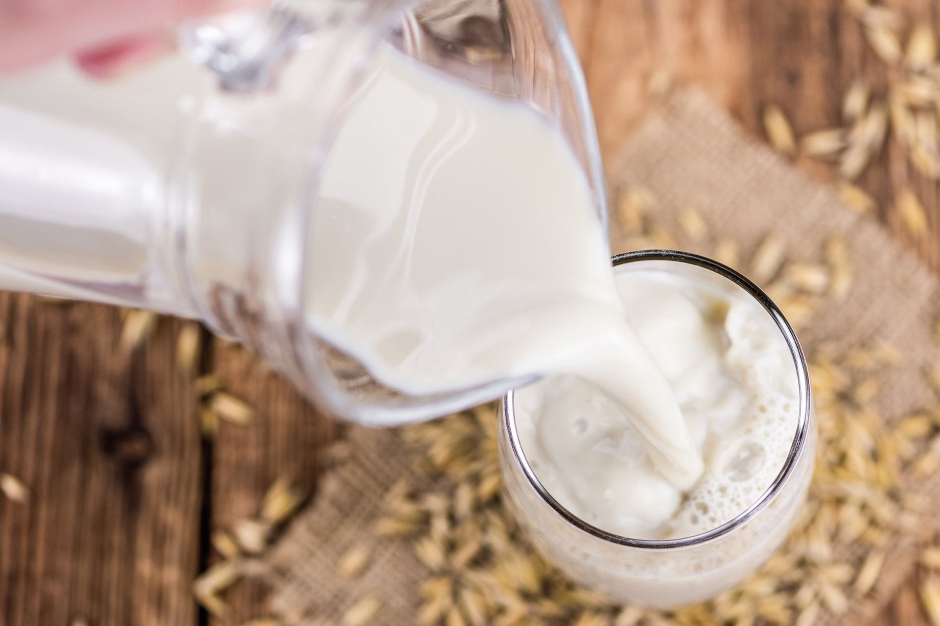 Milk being poured from a glass pitcher into a glass, surrounded by scattered grains on a wooden surface.