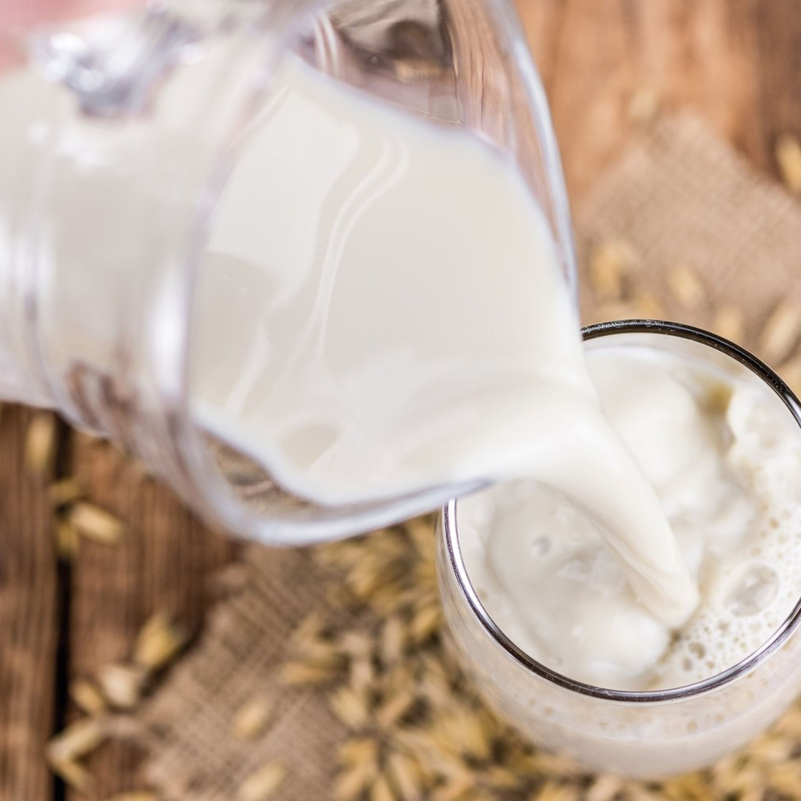 Milk being poured from a glass pitcher into a glass, surrounded by scattered grains on a wooden surface.