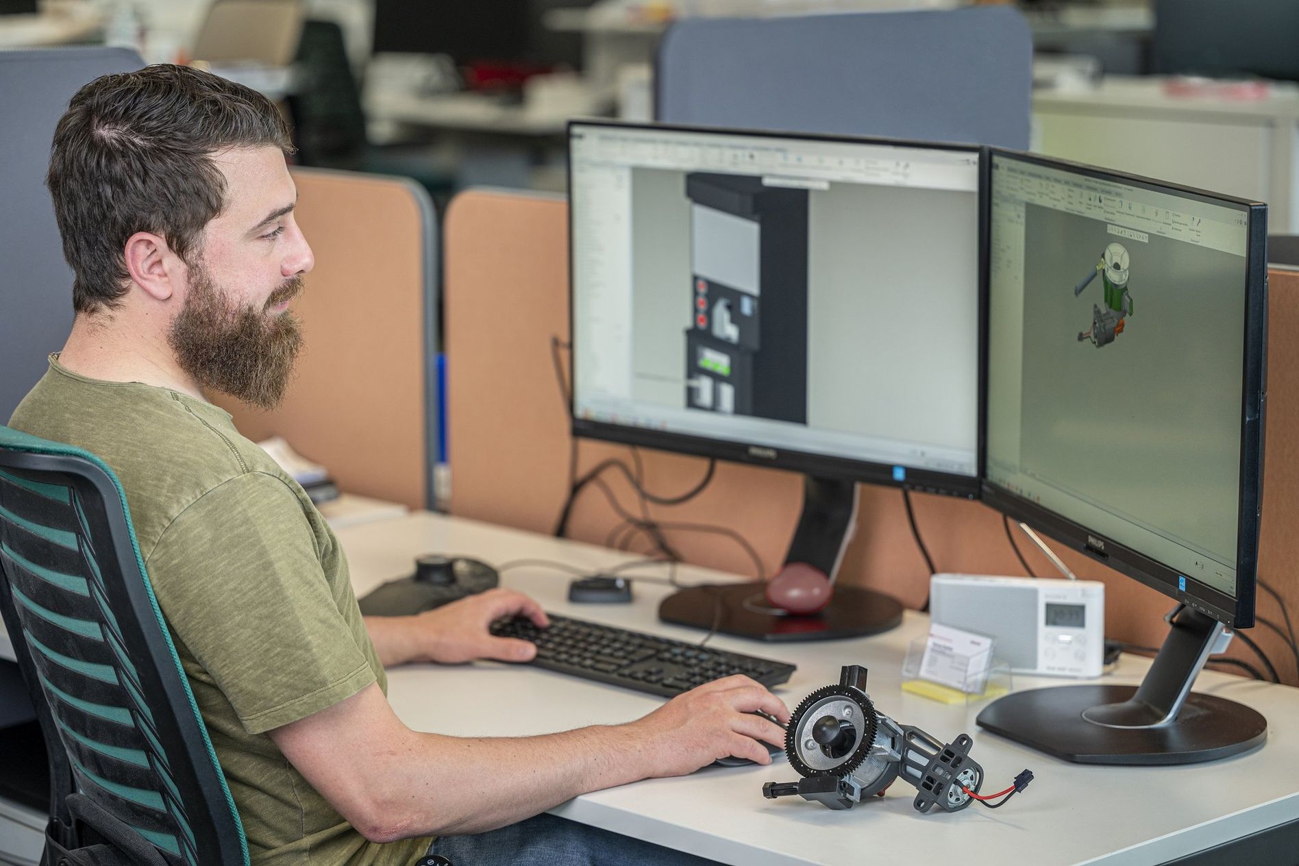 A man at a desk works on 3D modeling software on dual monitors, holding a mechanical part. Office environment in the background.