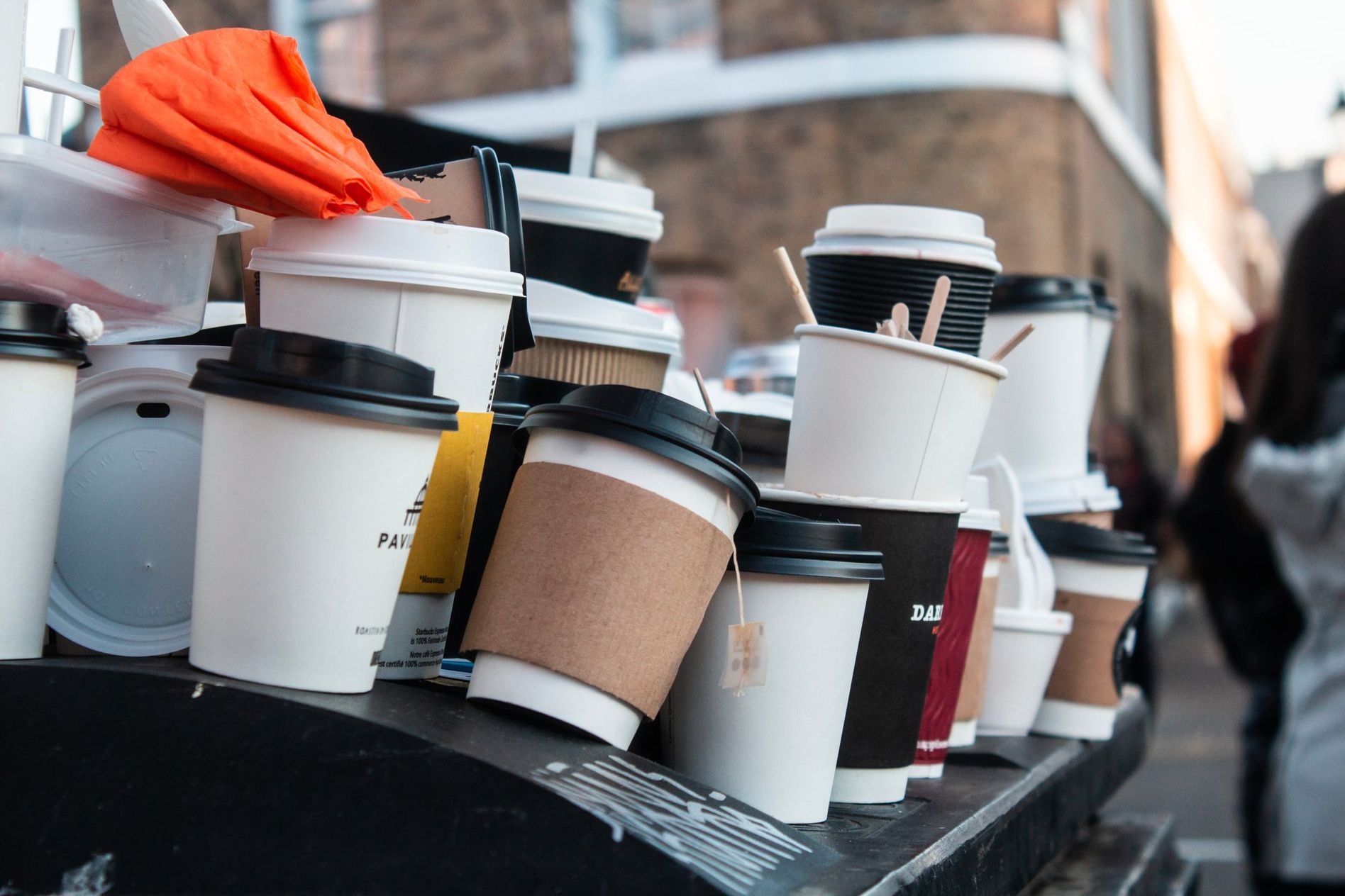 A pile of various disposable coffee cups, some with lids, stacked on an outdoor surface. A blurred street scene is in the background.
