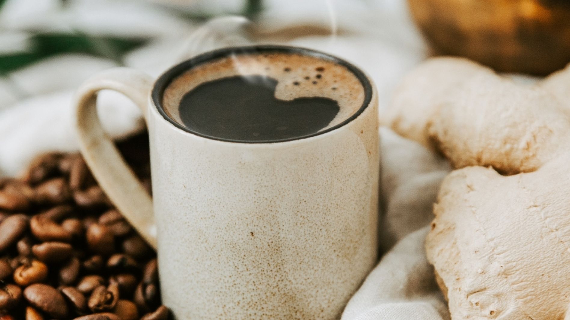 A ceramic mug of coffee sits next to scattered coffee beans and a large piece of ginger, with green leaves in the background.