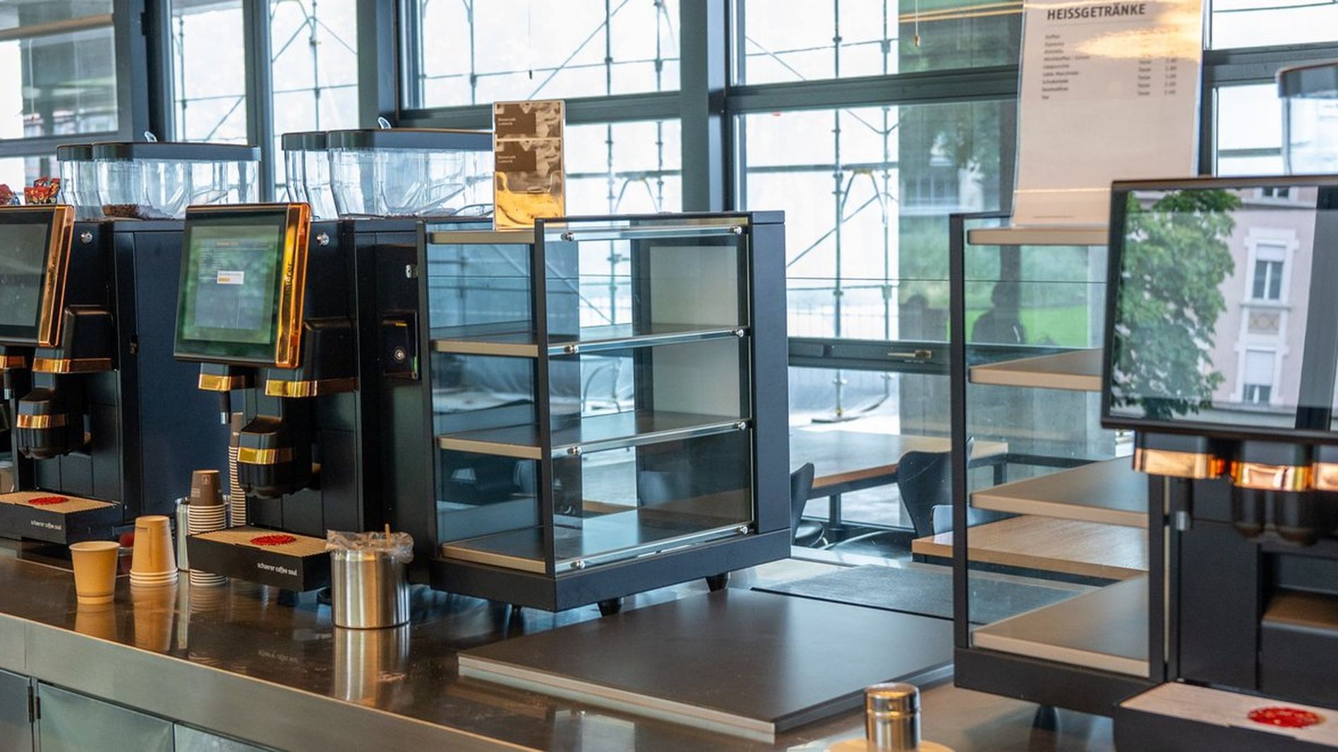 Modern self-service coffee machines and empty bakery display cases in a bright cafeteria with large windows.