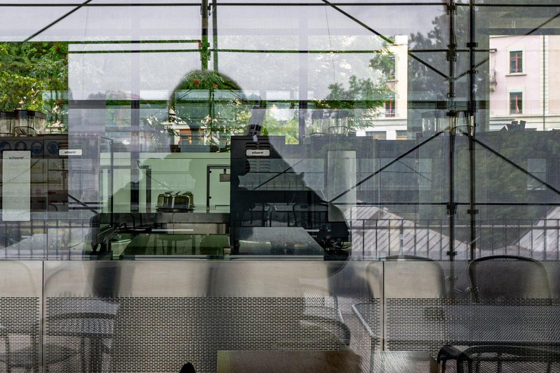 Reflection of a person taking a photo through a glass window, showing an empty café interior with tables and chairs, and a street view outside.