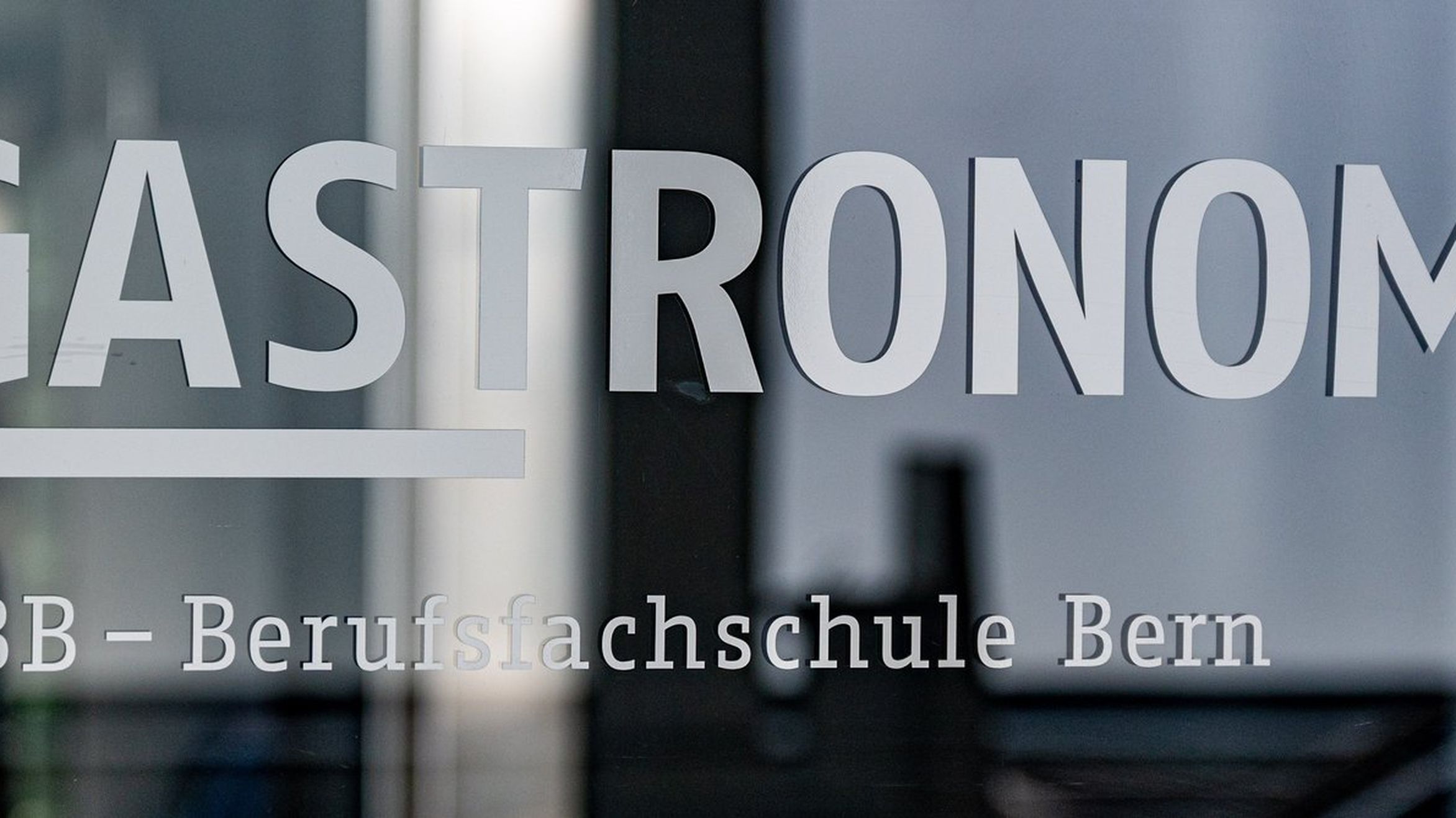 Glass door with "GASTRONOMIE" and "GIBB - Berufsfachschule Bern" written on it, reflecting a blurred interior scene.