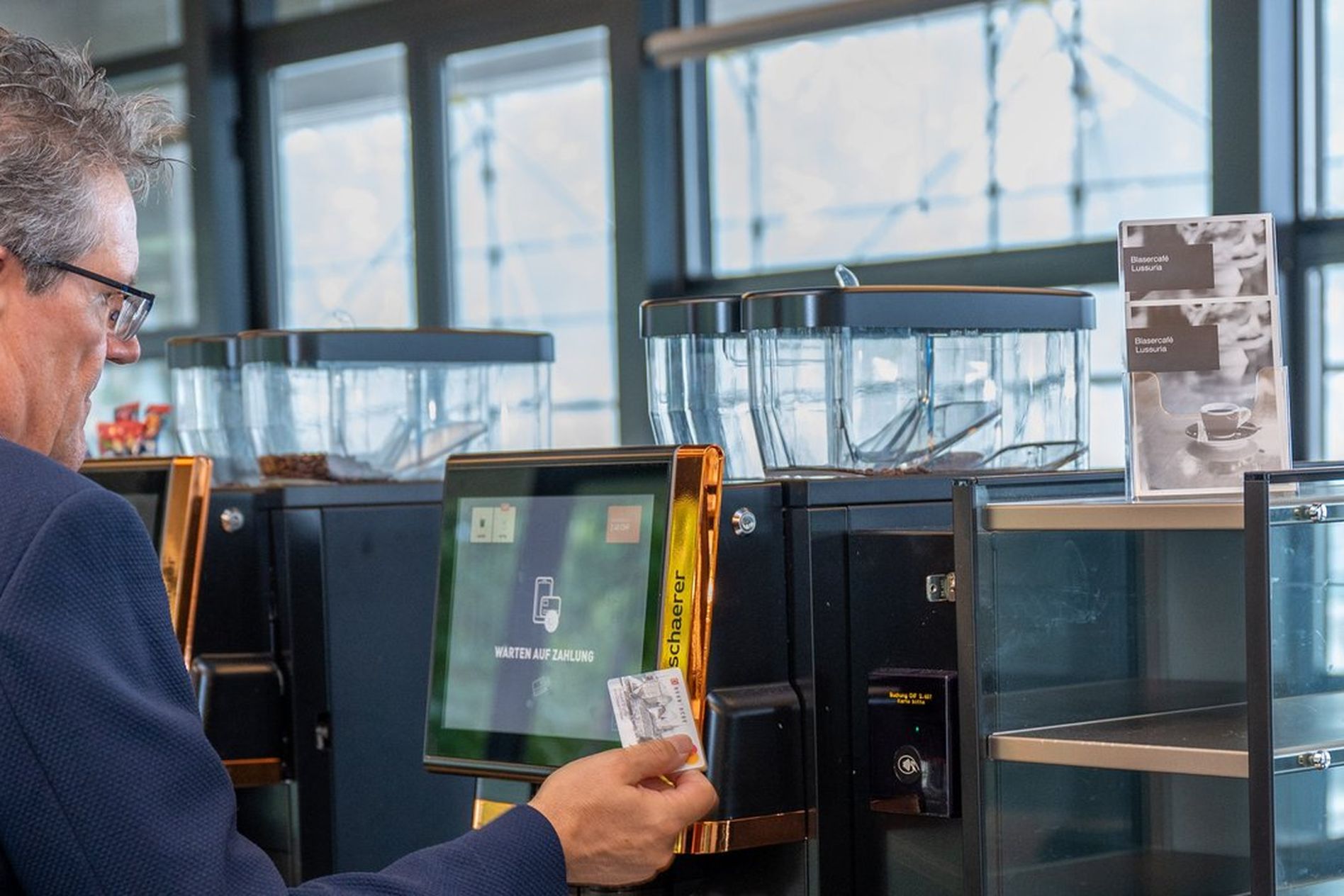 A man in a suit uses a touchscreen on a modern vending machine in a bright, glass-walled room.
