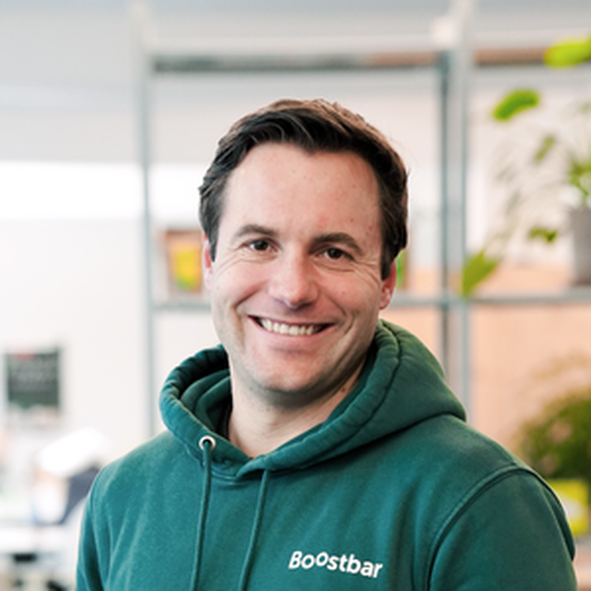 Smiling person in a green hoodie with "Boostbar" logo, standing indoors with blurred background.