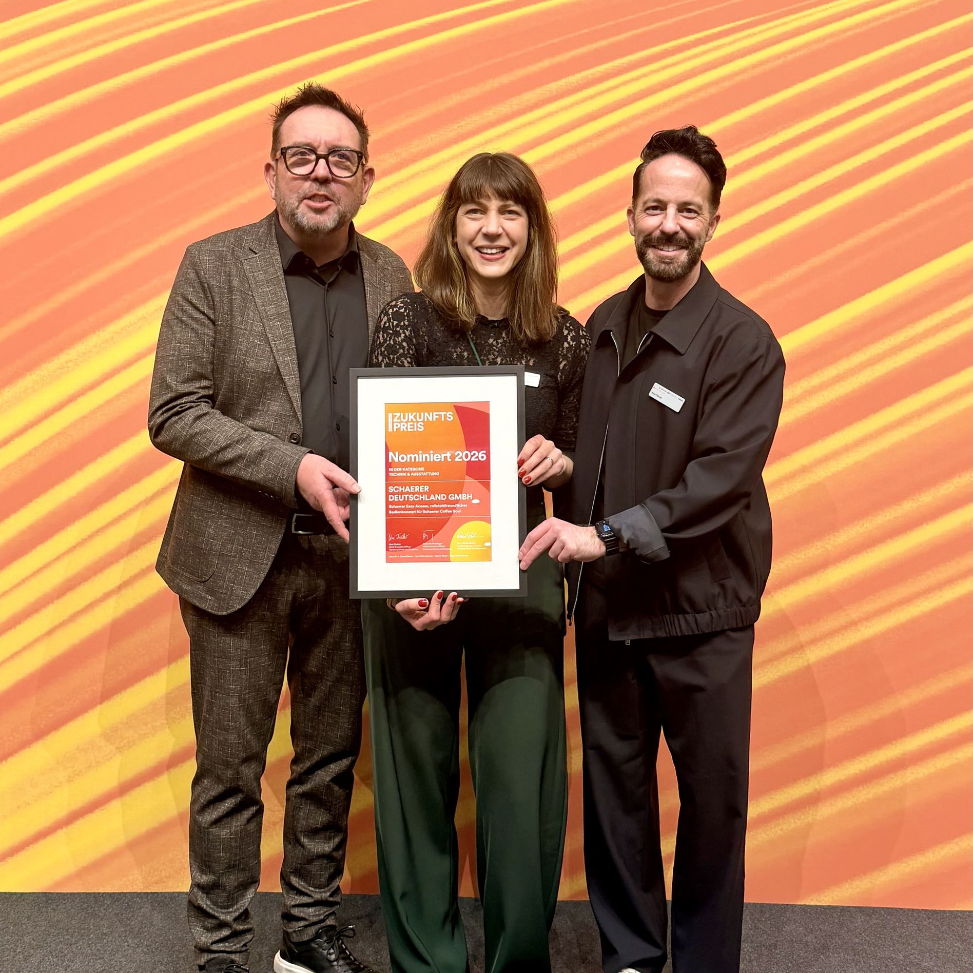 Three people smiling and holding an award certificate against an orange patterned background.