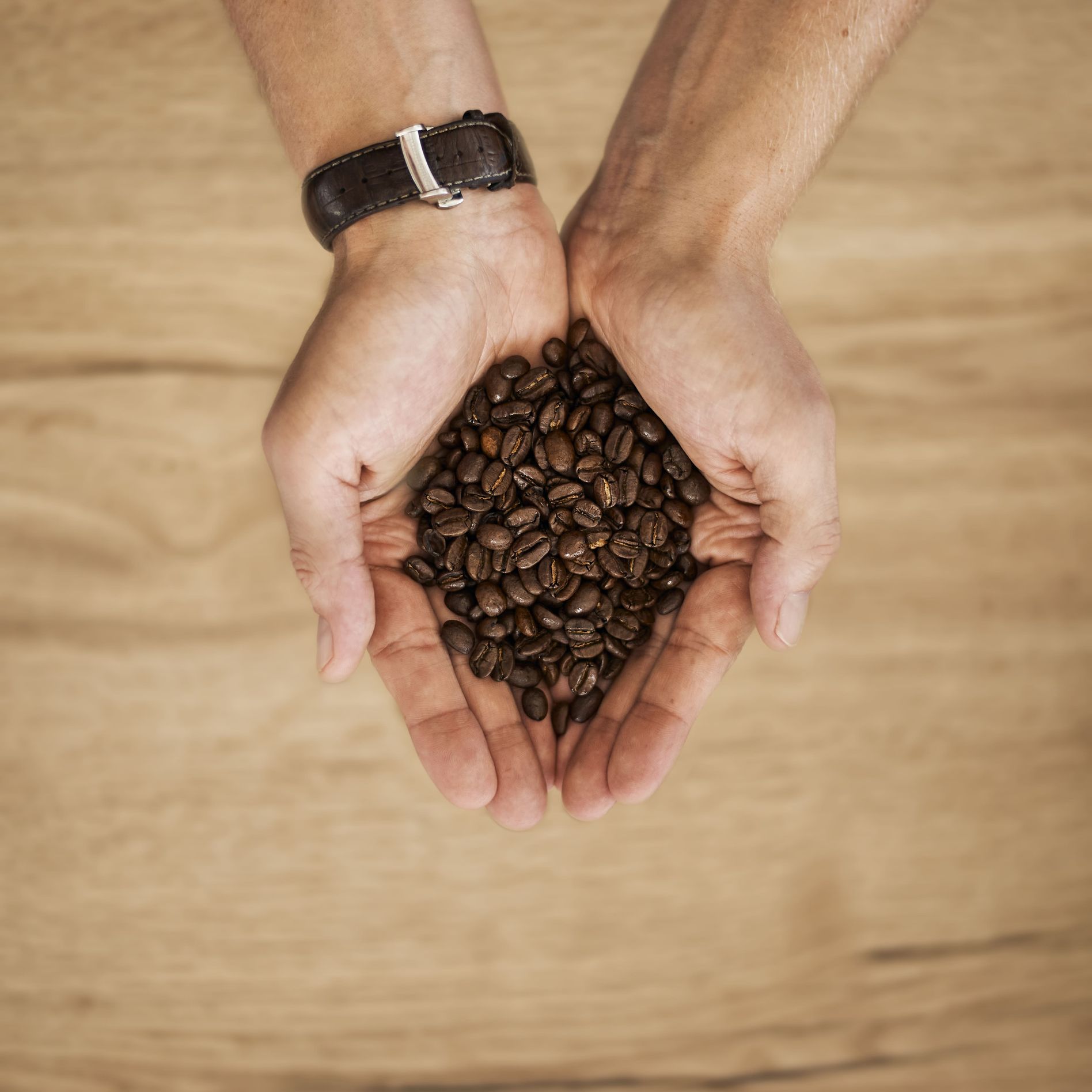 Hands holding roasted coffee beans over a wooden background.
