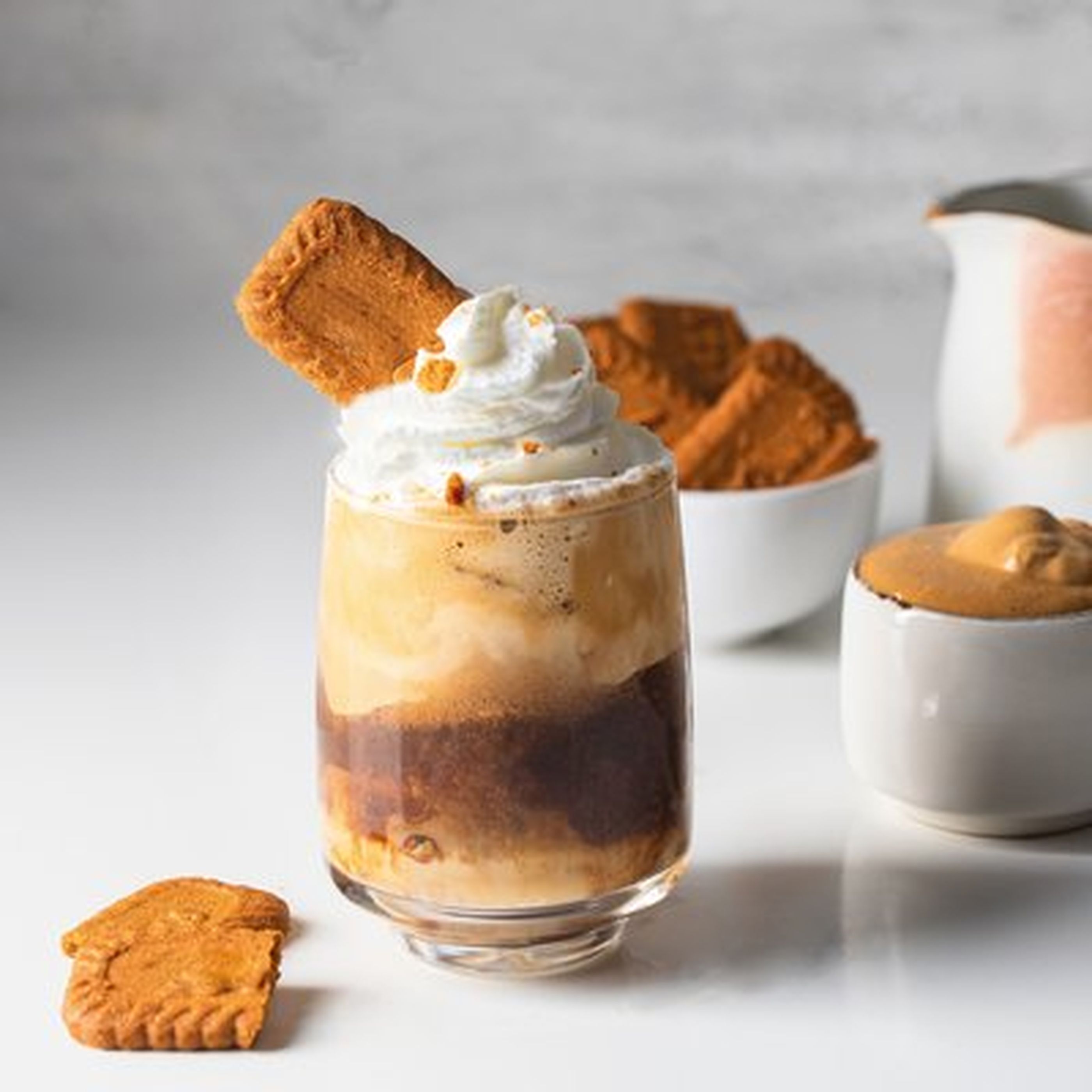 Glass of iced coffee topped with whipped cream and a cookie, surrounded by bowls of cookies and creamy spread on a white surface.