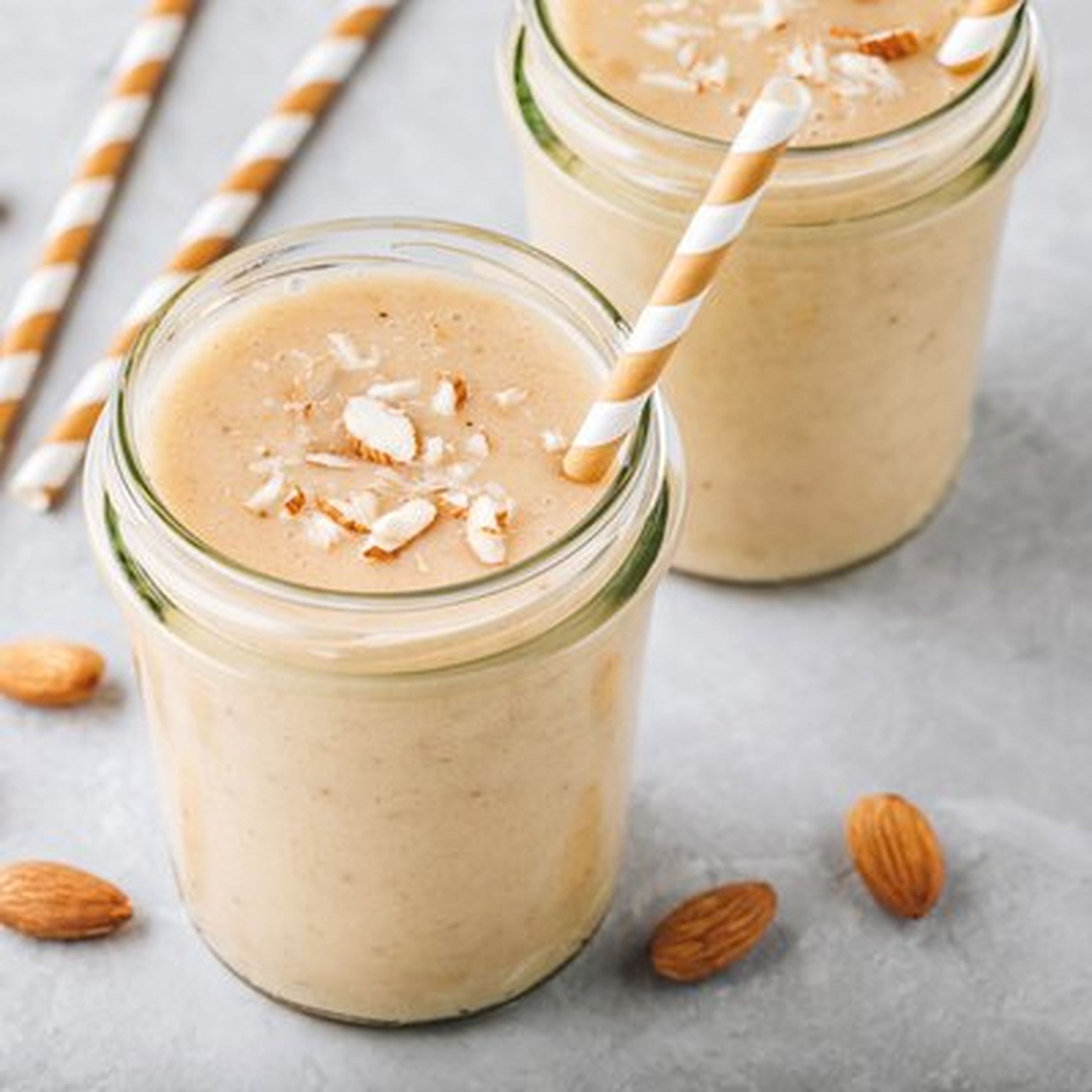 Two glasses of creamy almond smoothie with striped straws, topped with almond slices, surrounded by whole almonds on a gray surface.