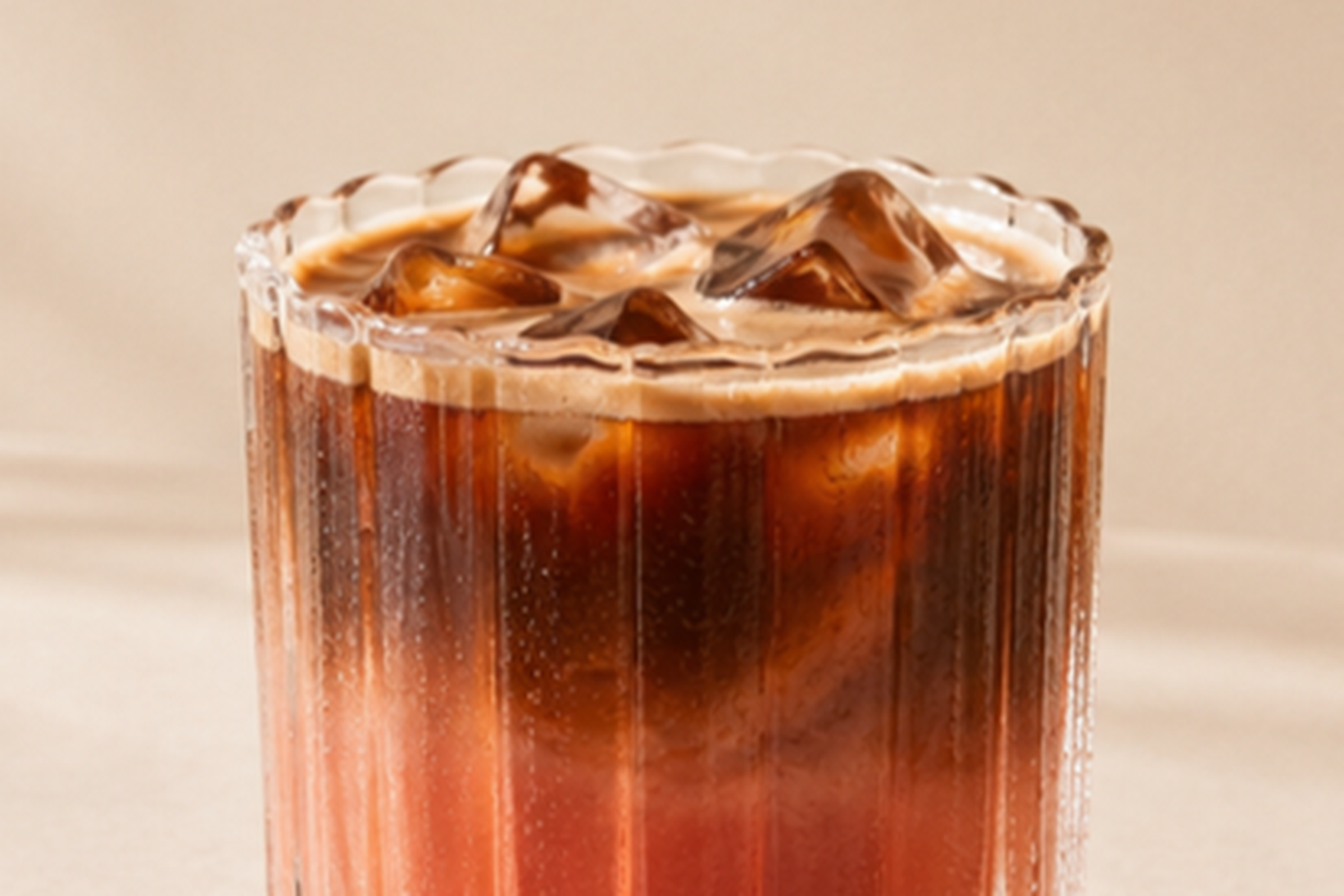 A glass of iced coffee with a creamy swirl on top, served in a ribbed tumbler against a neutral background.
