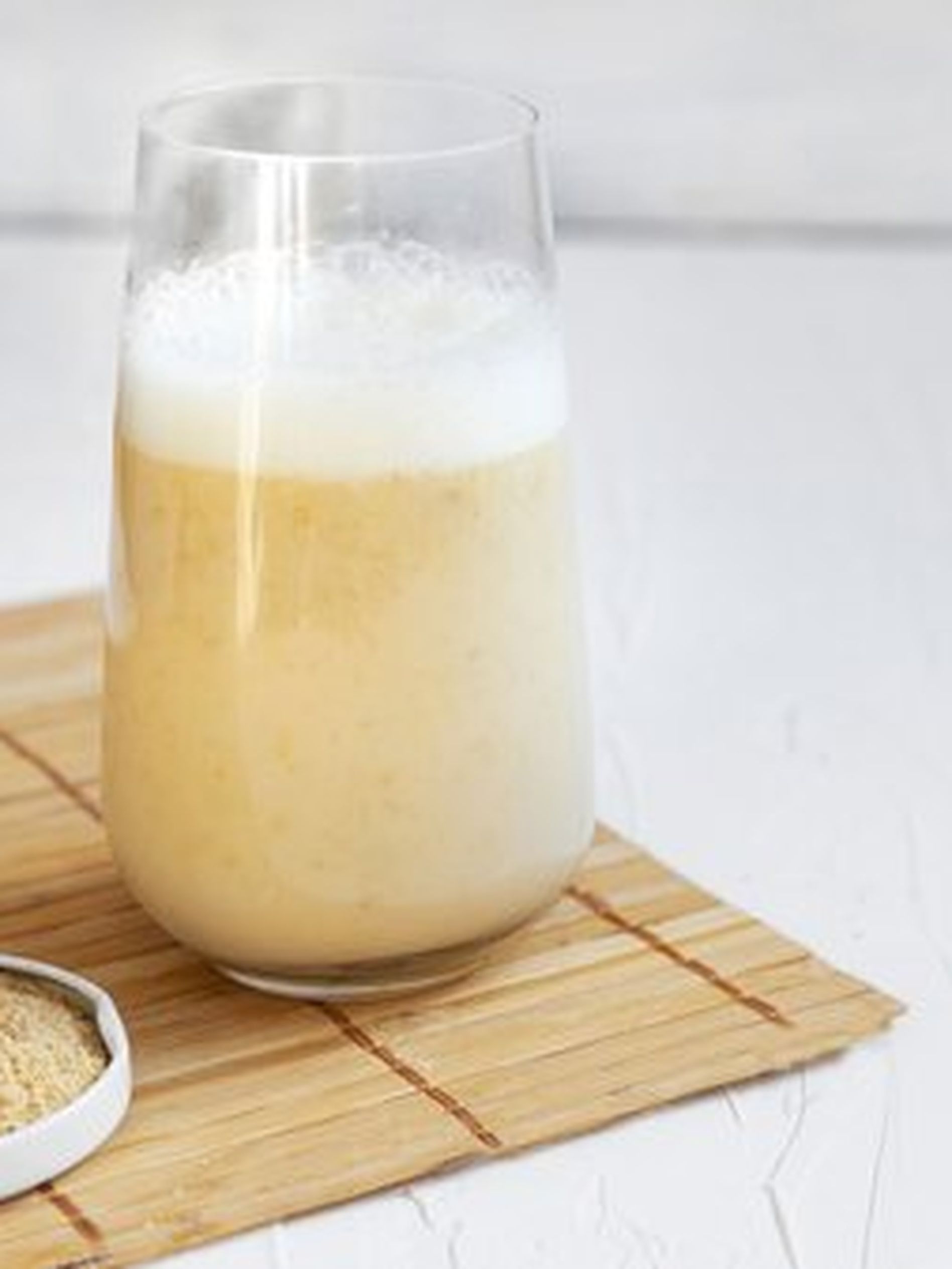 A glass of frothy beige smoothie on a bamboo mat, with a small dish of powder beside it.