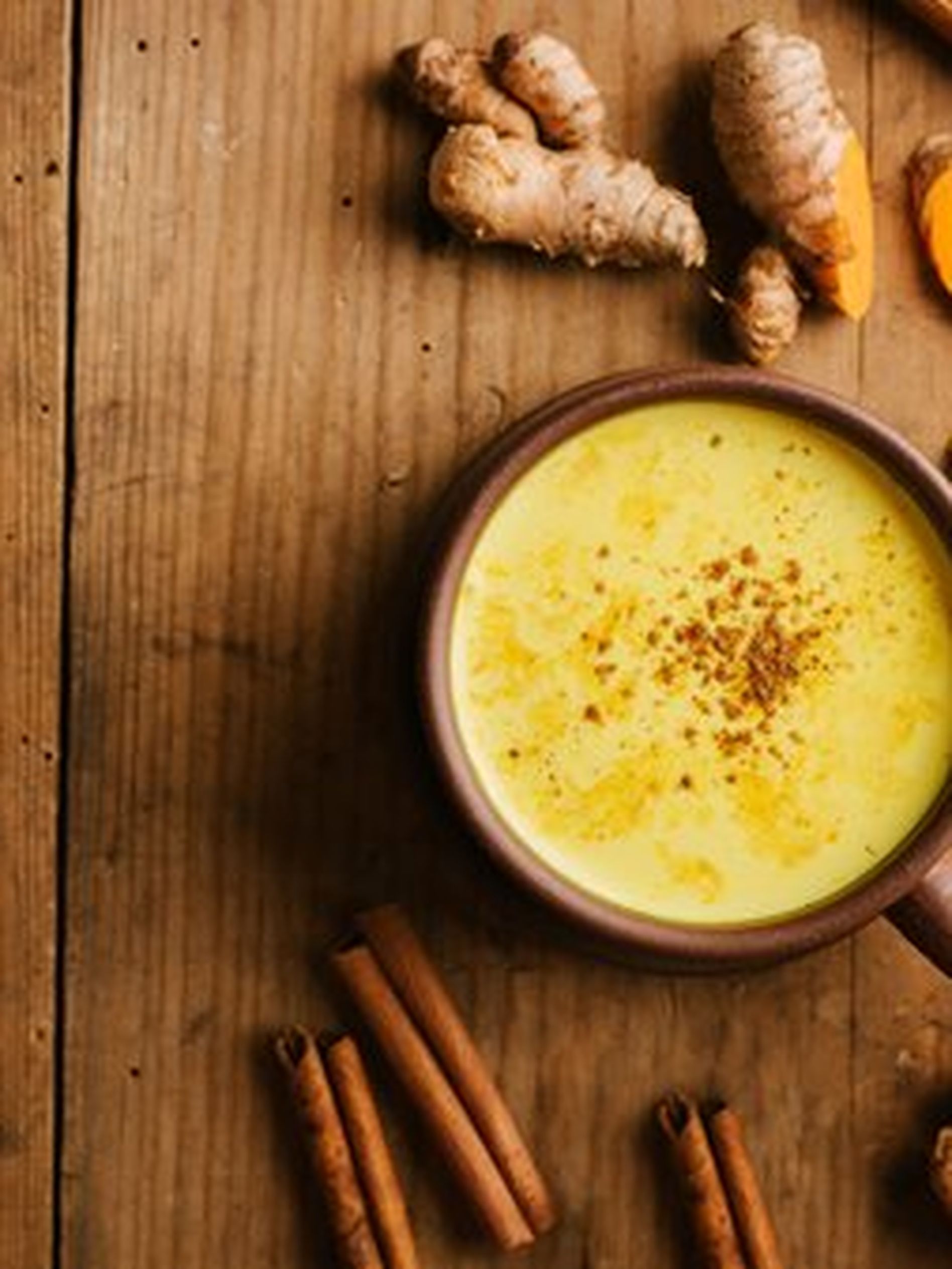 A cup of golden turmeric milk on a wooden table, surrounded by turmeric roots, powder, and cinnamon sticks.