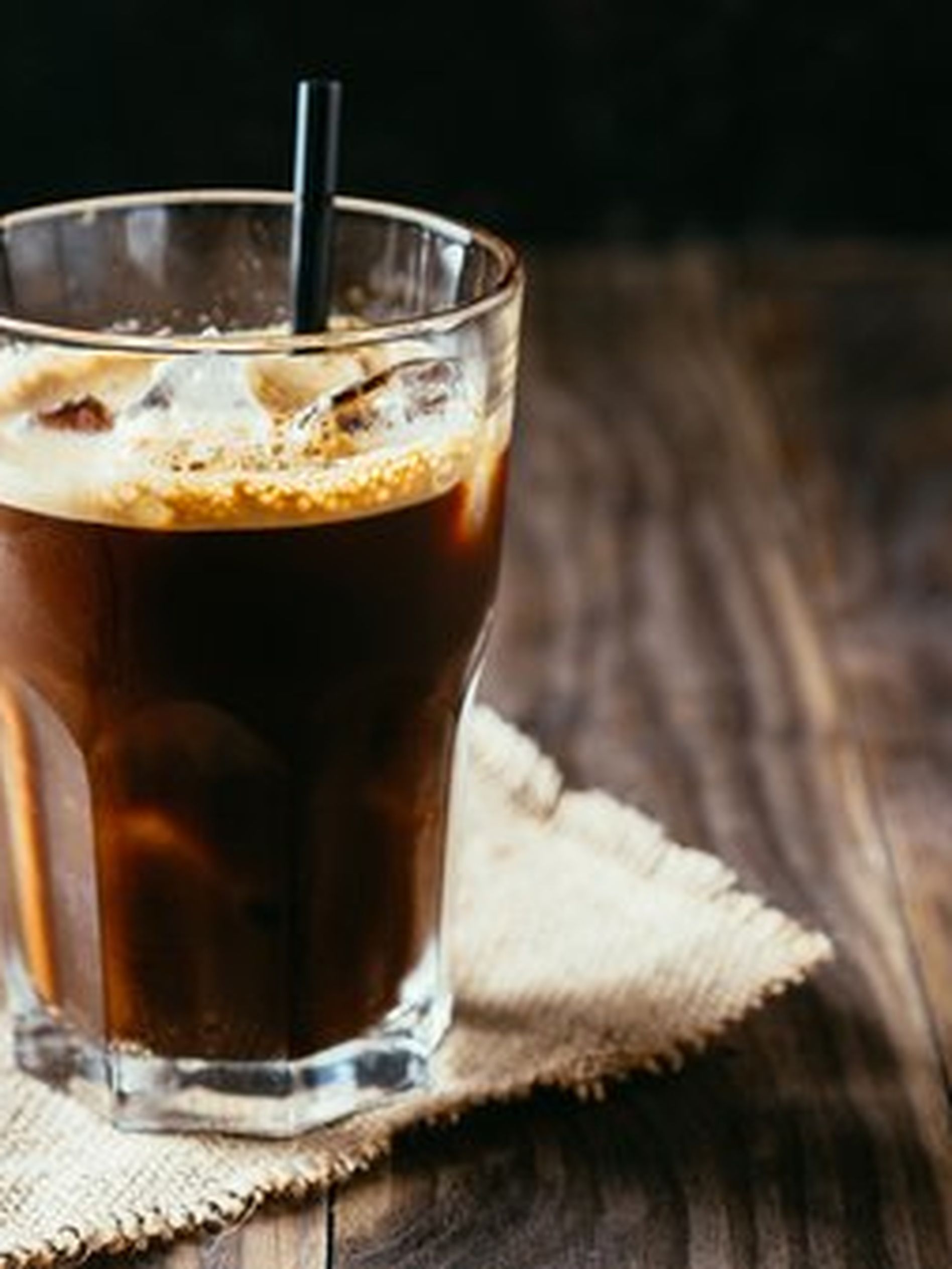 A glass of iced coffee with a black straw on a burlap cloth, set on a wooden table with a dark background.