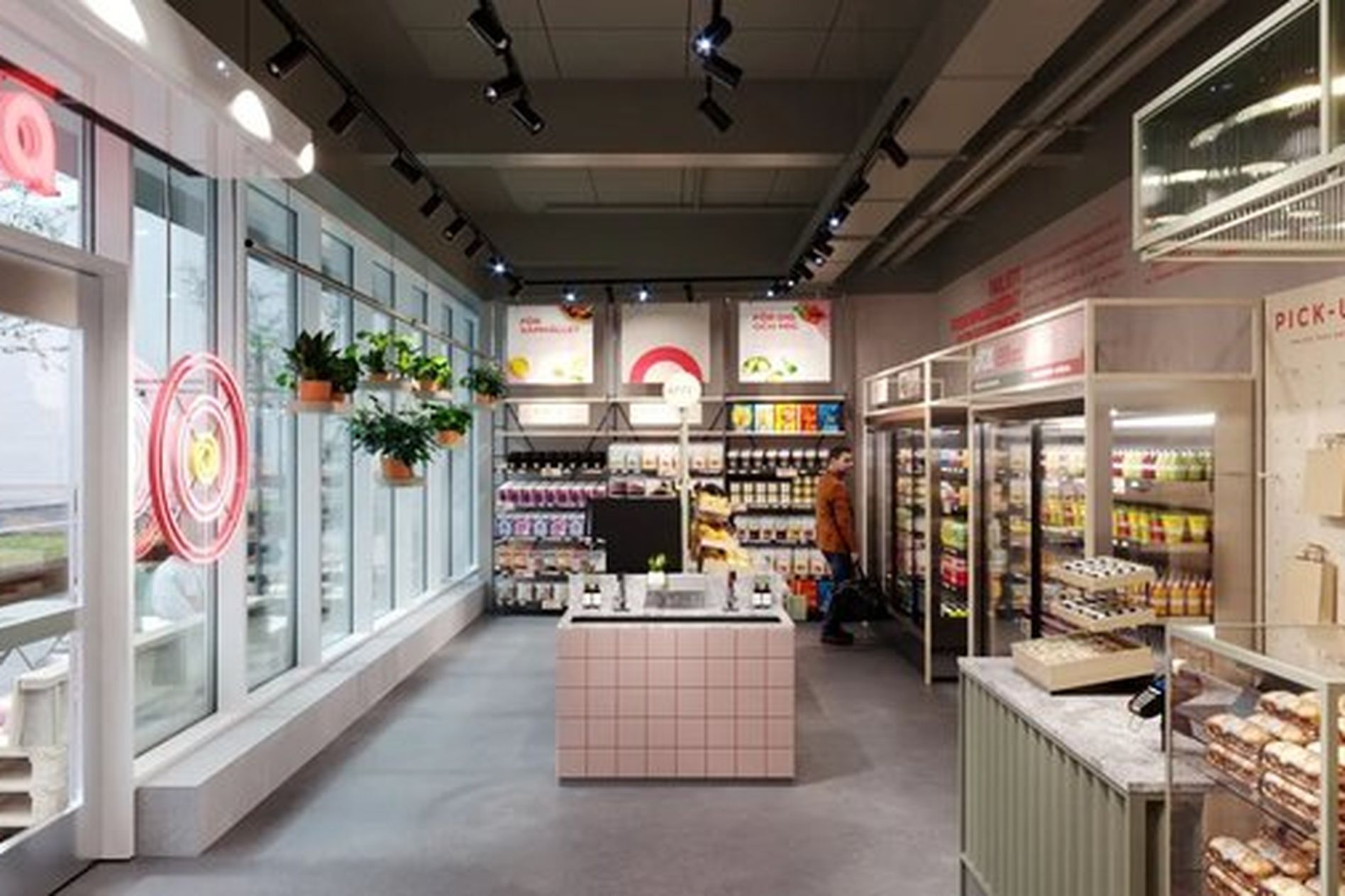 Modern convenience store interior with large windows, shelves stocked with various products, a pick-up area, and hanging plants.