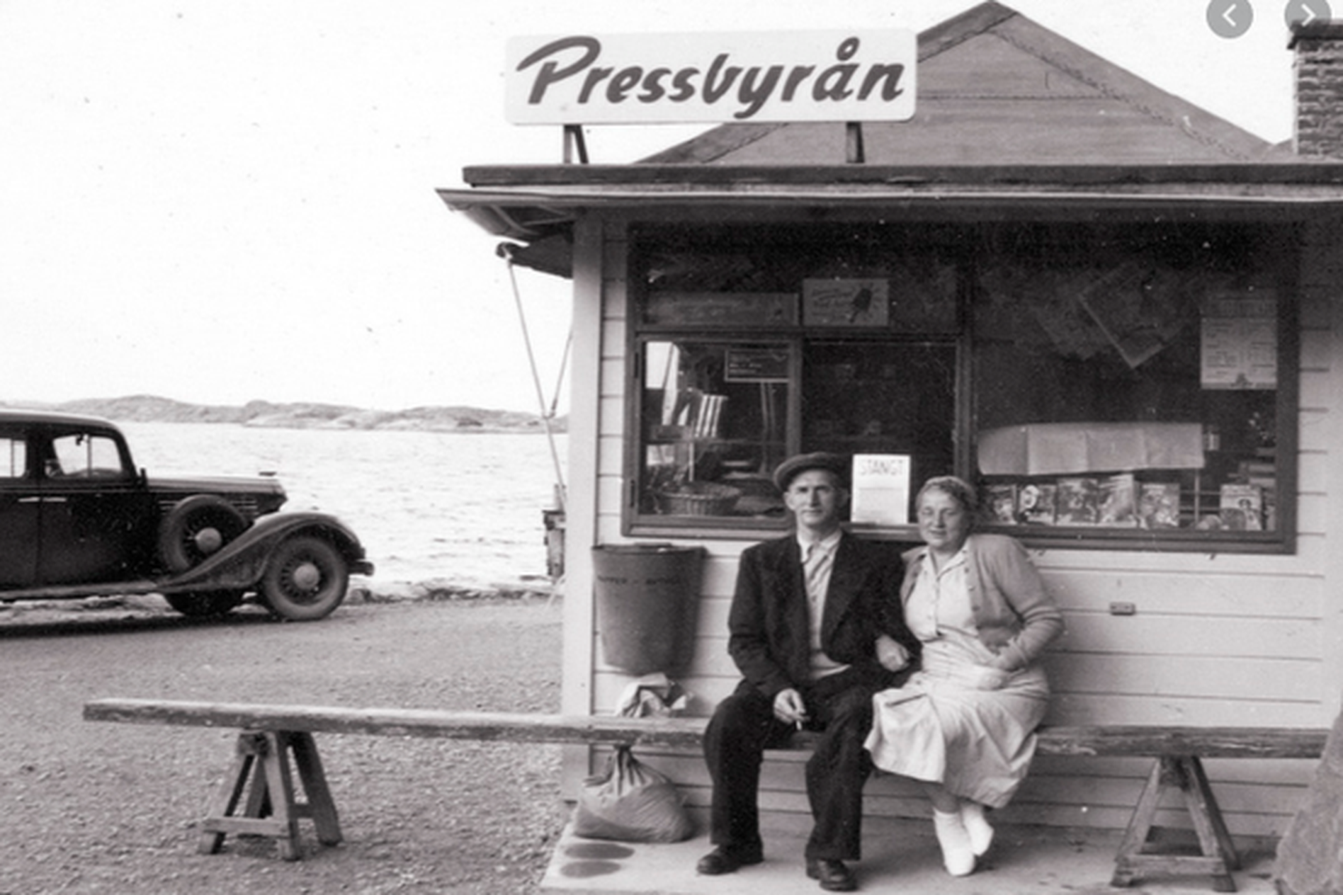 A vintage photo of a man and woman sitting on a bench outside a small kiosk labeled "Pressbyrån," with a classic car and sea in the background.