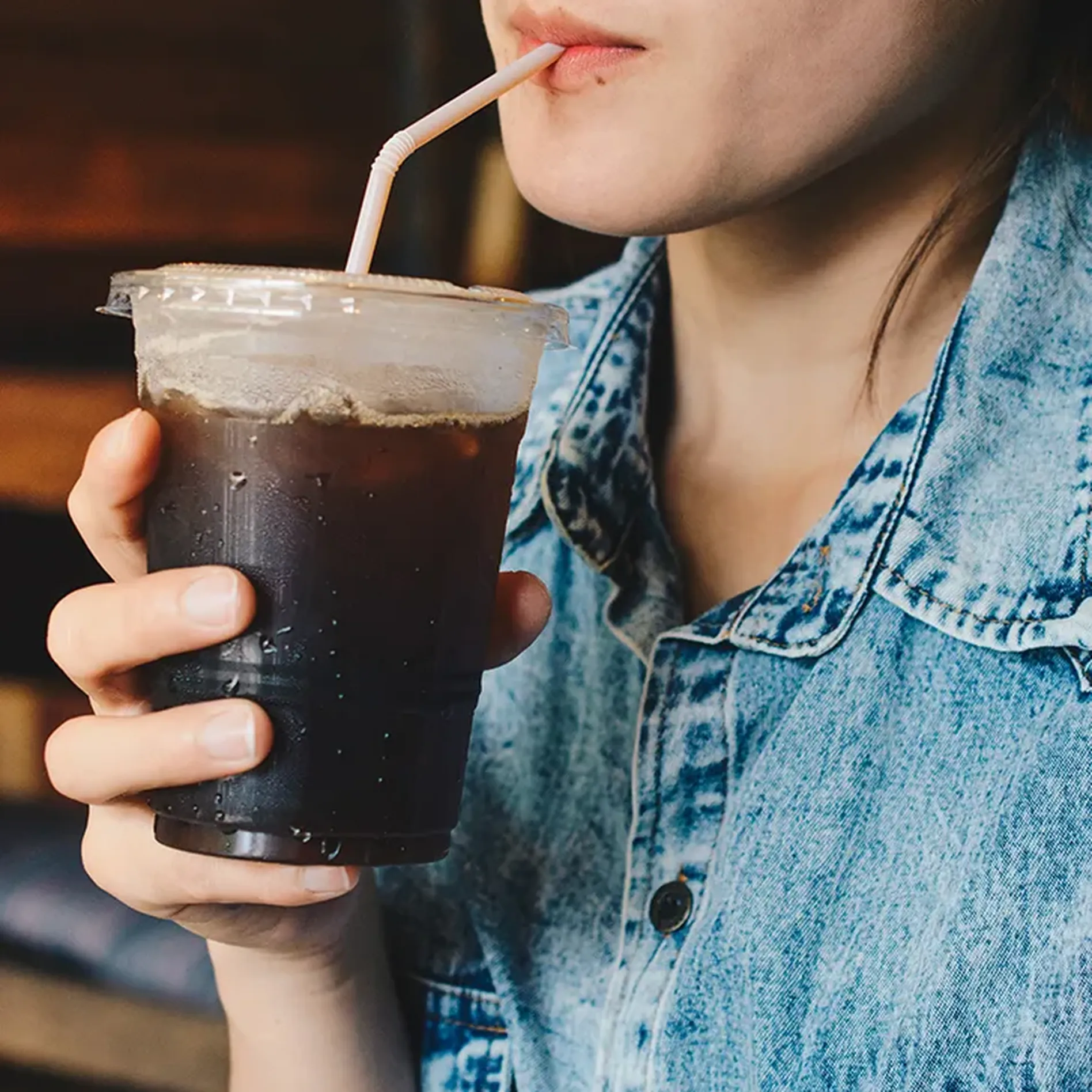Person in a denim shirt drinking iced coffee from a plastic cup with a straw.