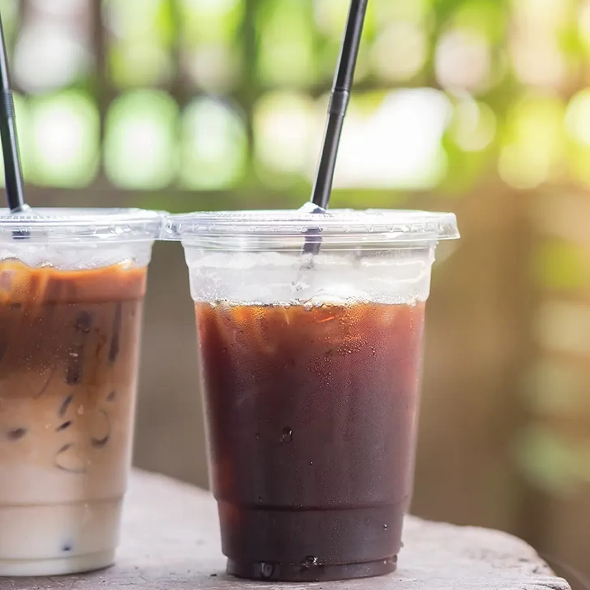 Two plastic cups with iced coffee; one is dark and the other is lighter with milk. They have black straws and sit on a wooden surface.